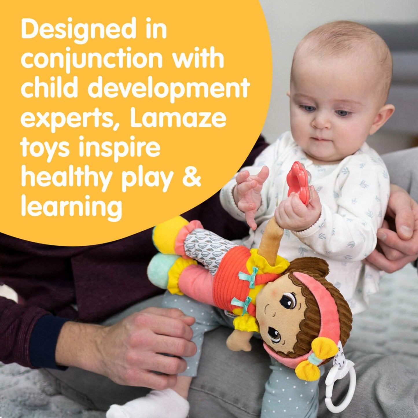 Baby sitting on parent’s lap, holding a Lamaze doll and red teether while text mentions developmental design with child experts to encourage learning.