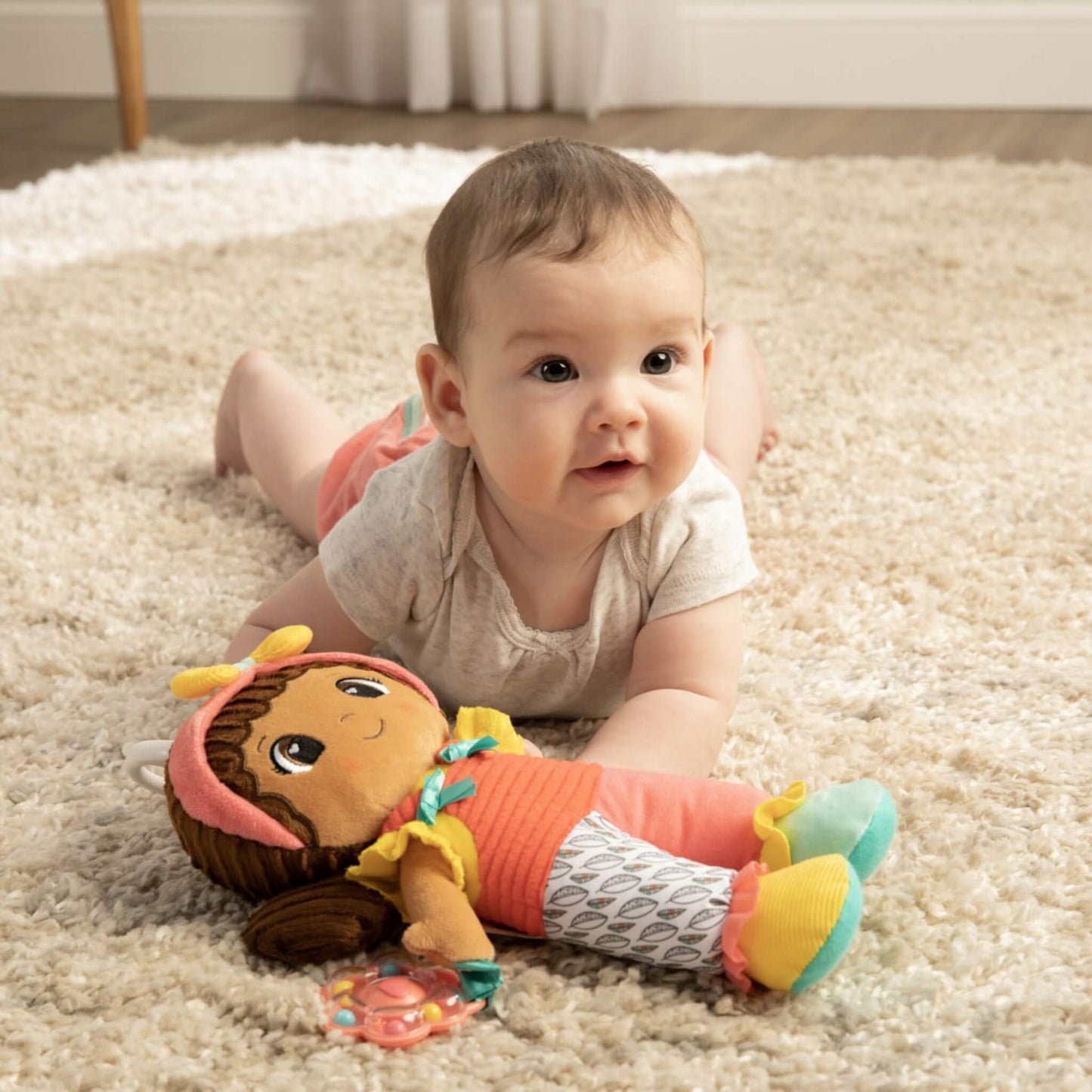 Baby lying on tummy on a soft rug, smiling while playing with a colourful doll dressed in red and yellow with a flower rattle nearby.