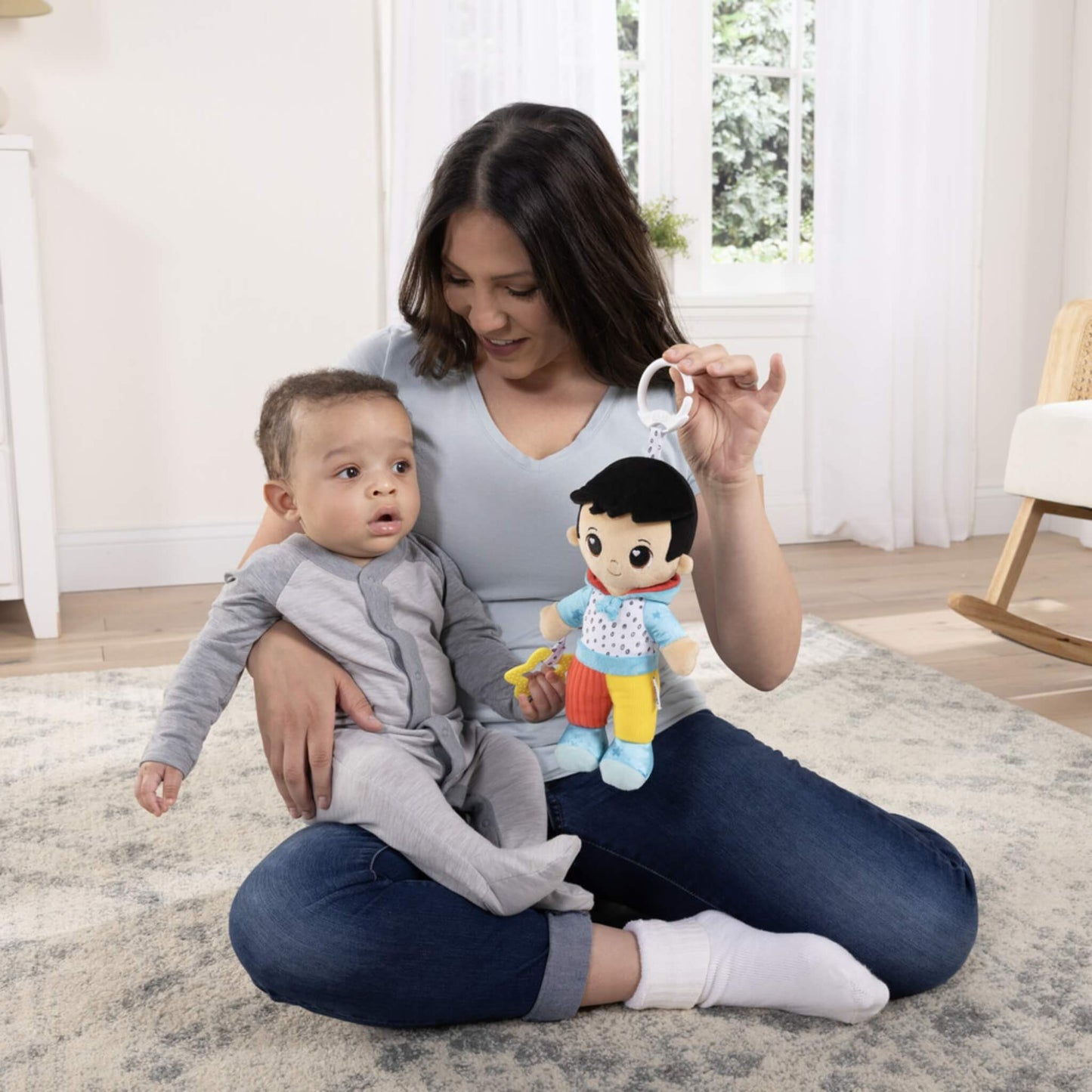 Mother sitting cross-legged on a soft rug holding baby on her lap while showing a soft doll with bright colours and a hanging clip.