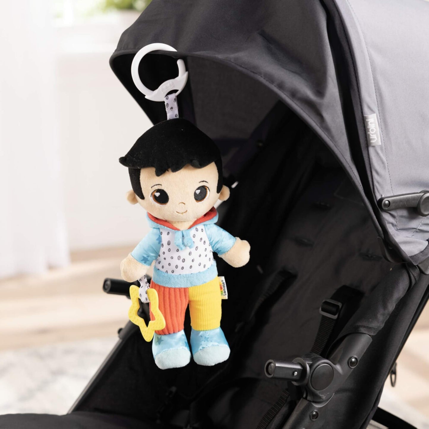 Soft doll hanging from a black stroller canopy using its clip, showing how it can attach securely for travel or outdoor walks.