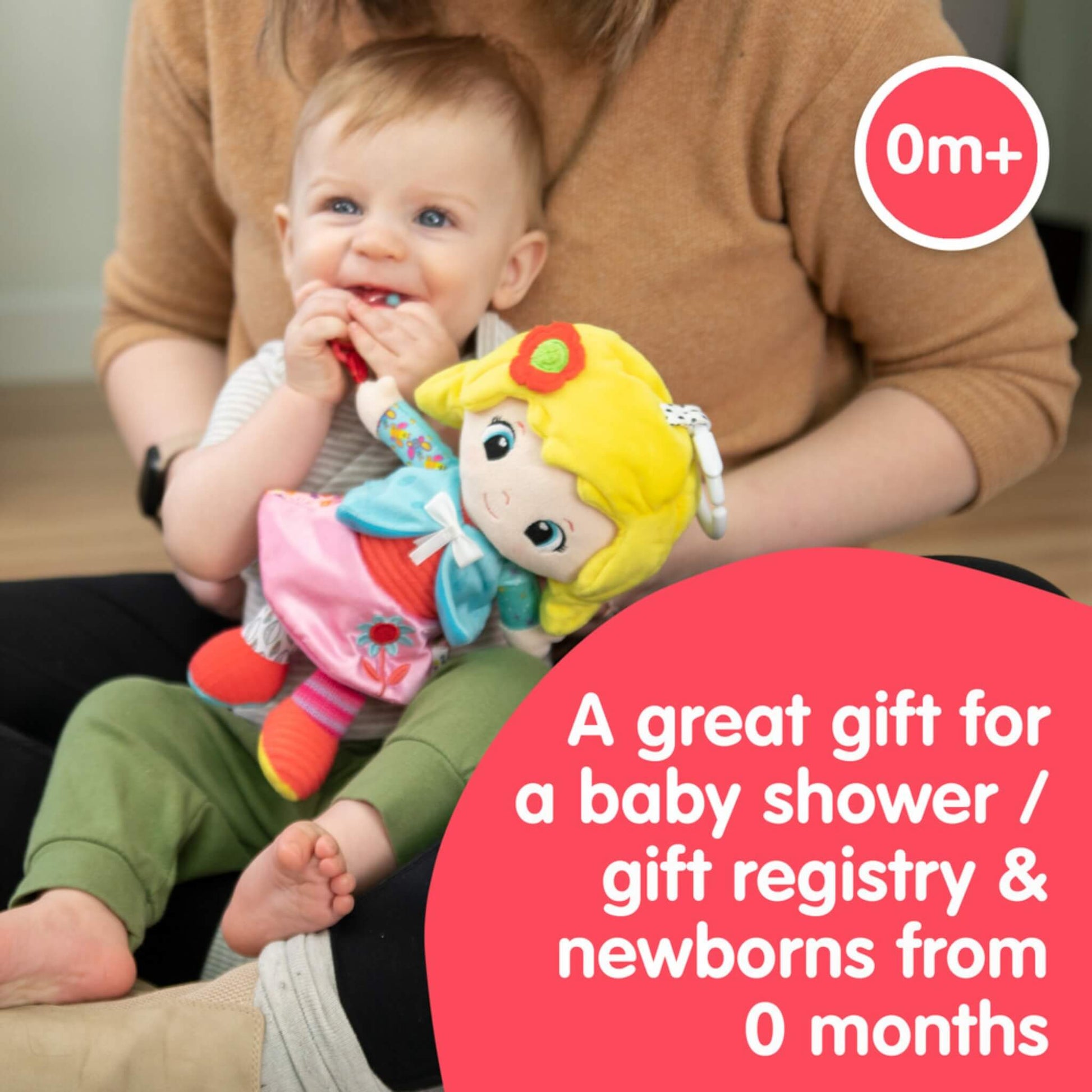 Baby sitting on a parent’s lap smiling and chewing on a flower-shaped rattle attached to a colourful doll with a blue jacket and pink skirt.