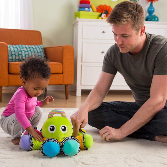 Baby sits on the floor beside an adult, exploring a bright green plush octopus toy together in a softly lit living room.
