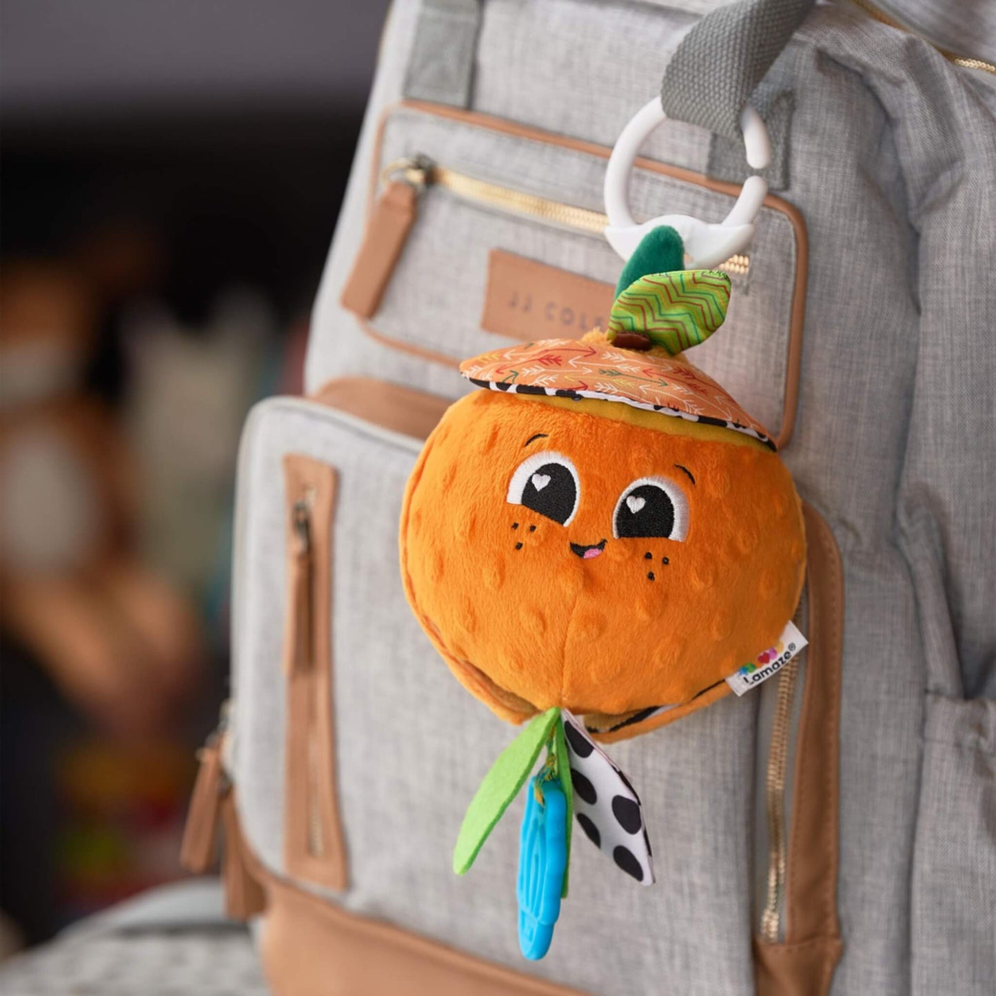 Soft orange-shaped baby toy with a smiling face, hanging from a light grey changing bag by a white clip, showing its portable on-the-go design.