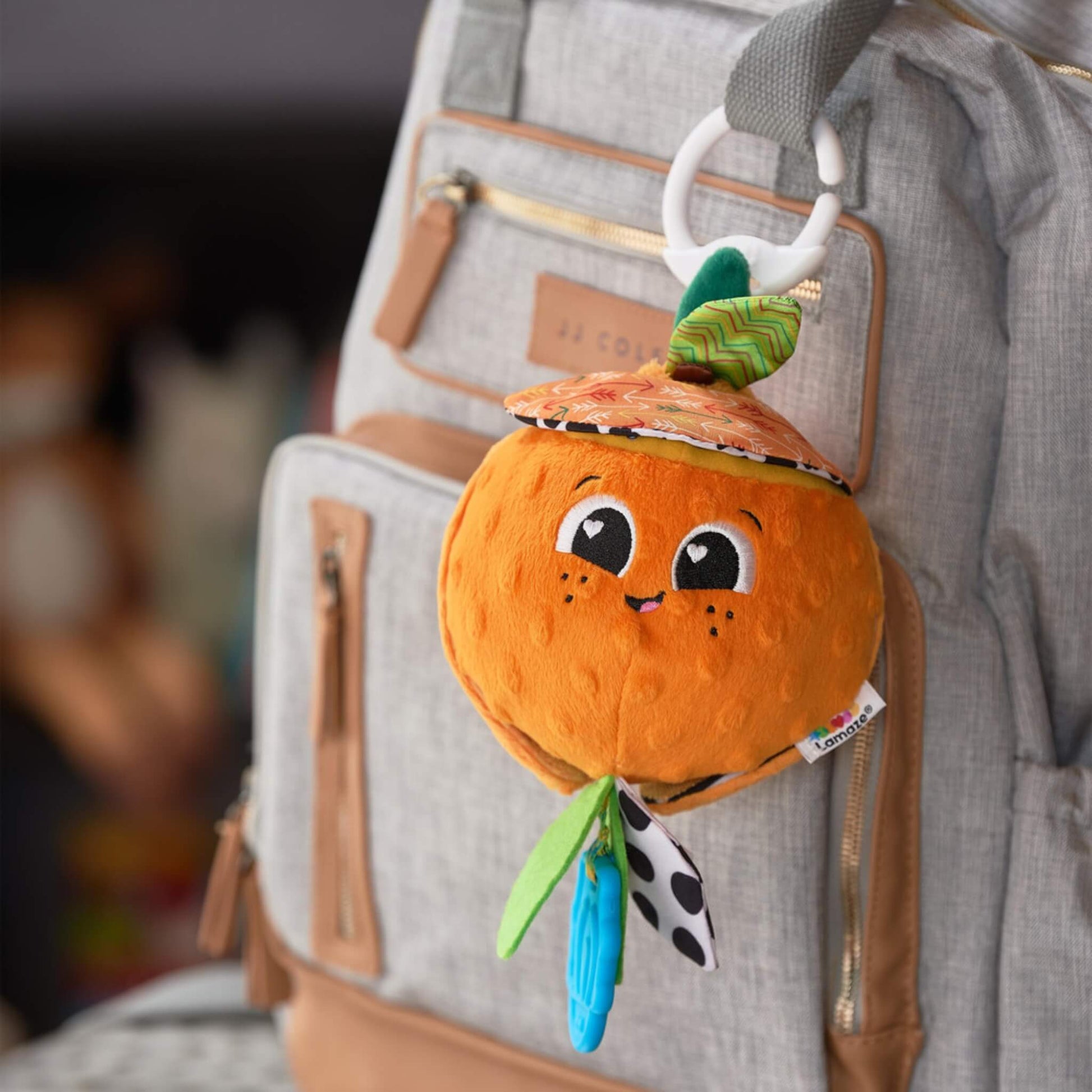Soft orange-shaped baby toy with a smiling face, hanging from a light grey changing bag by a white clip, showing its portable on-the-go design.