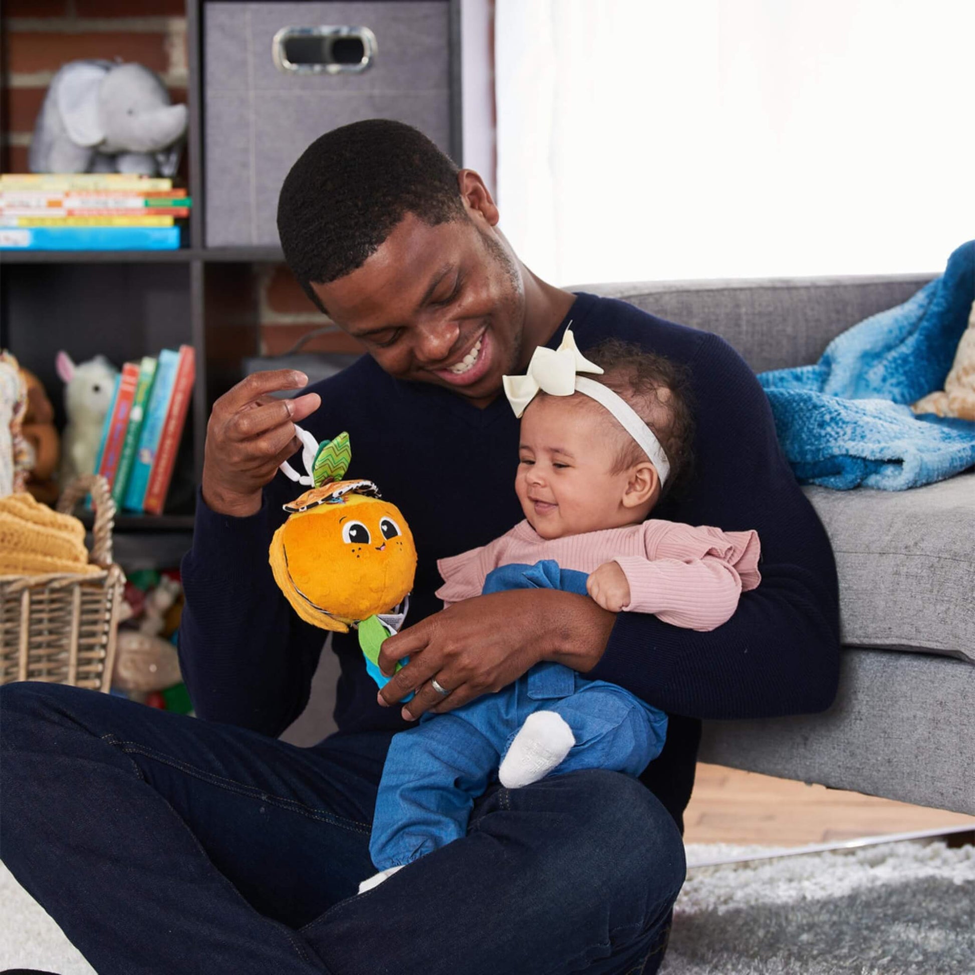 Baby sitting on an adult’s lap, reaching out to play with a plush orange toy featuring textured fabric, green leaves, and a bright smiling face.