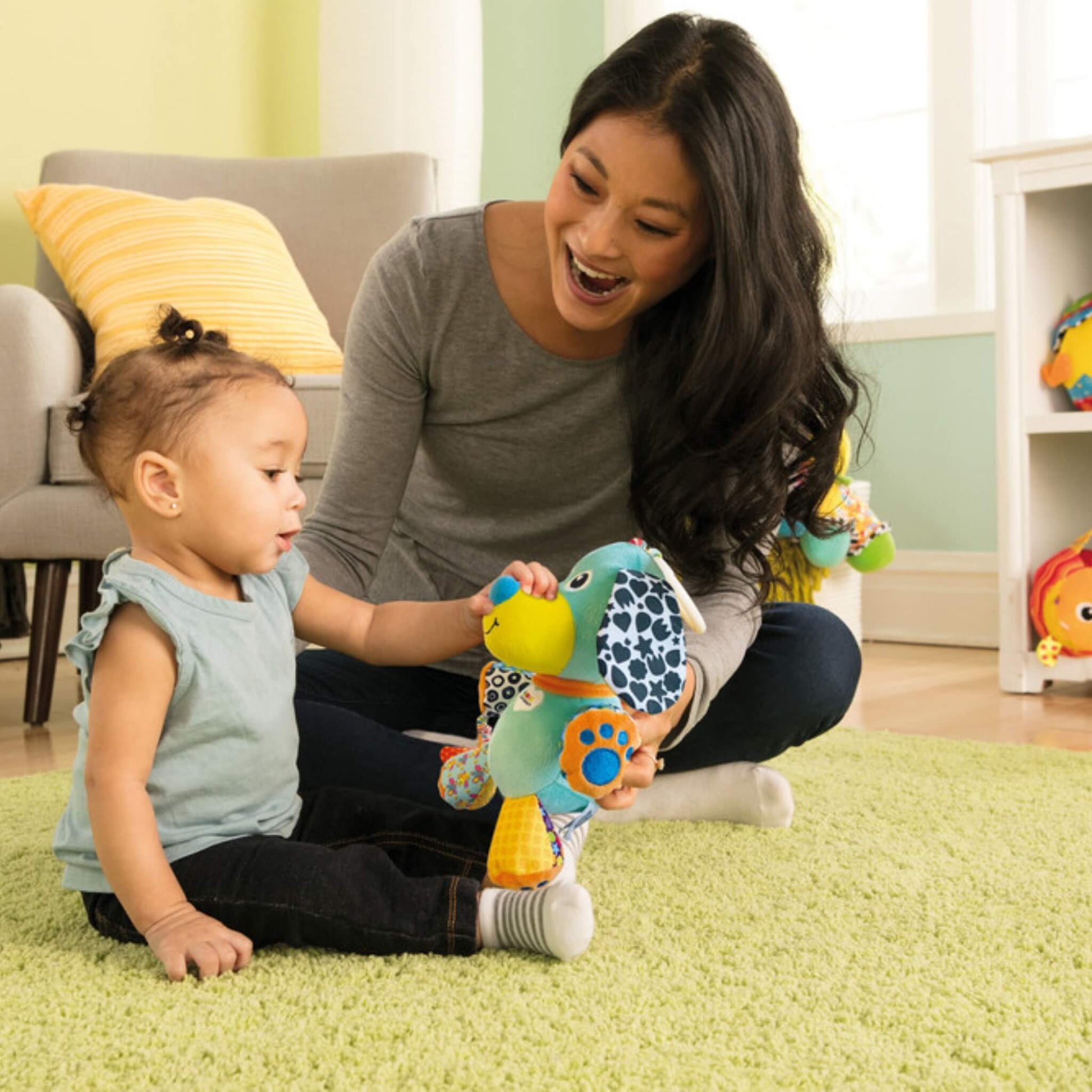 Baby sitting on a green rug exploring the Pupsqueak toy with their mother during playtime.