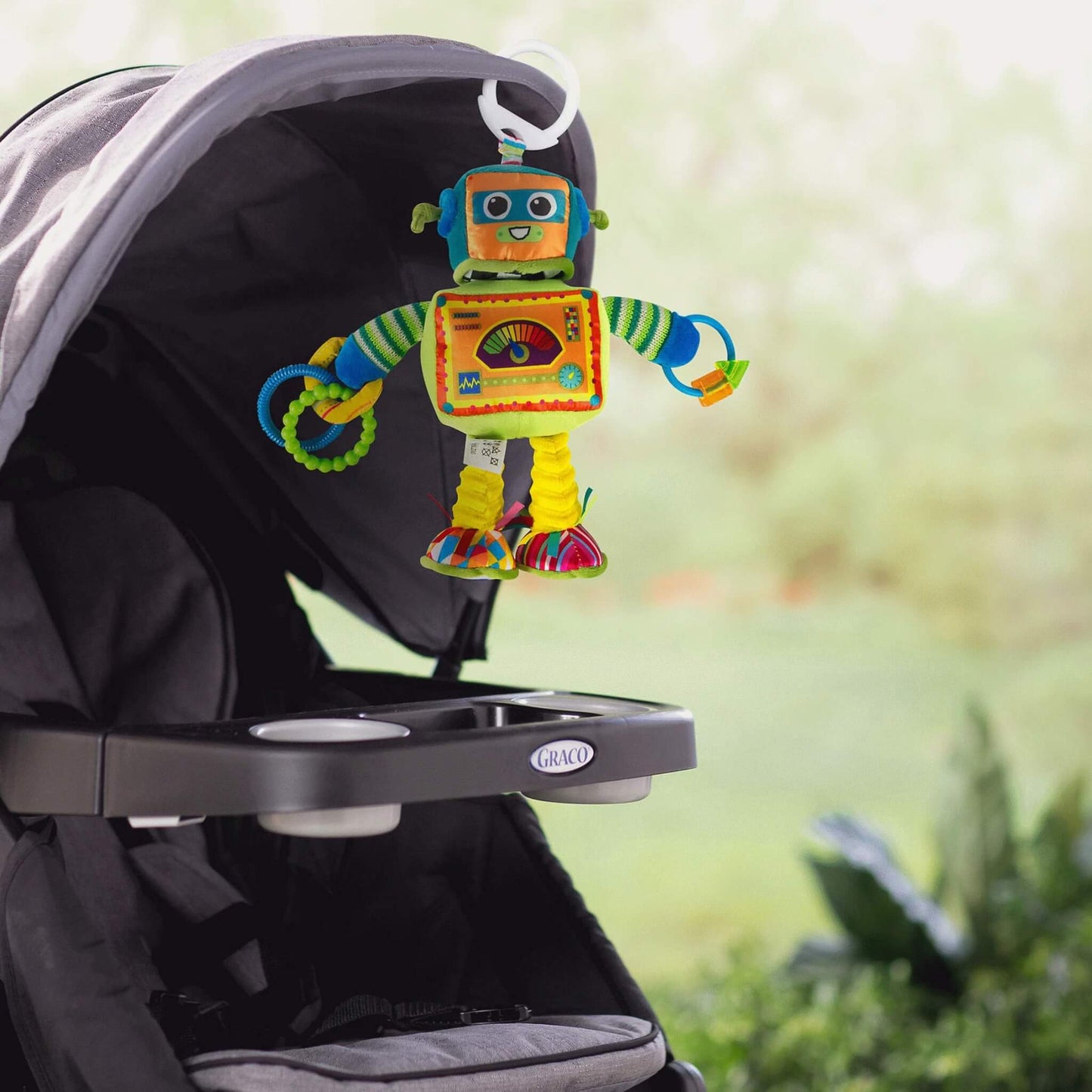 Soft robot toy clipped to a pram canopy outdoors, hanging above the seat where a baby can reach and interact during walks or playtime outside.
