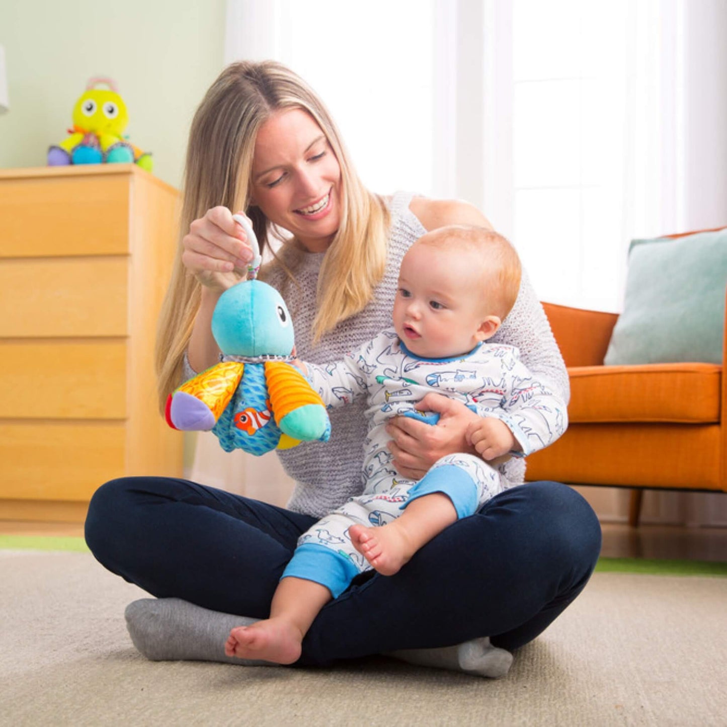 Mother sitting cross-legged on the floor, holding her baby and dangling a soft octopus toy for visual engagement and bonding play.