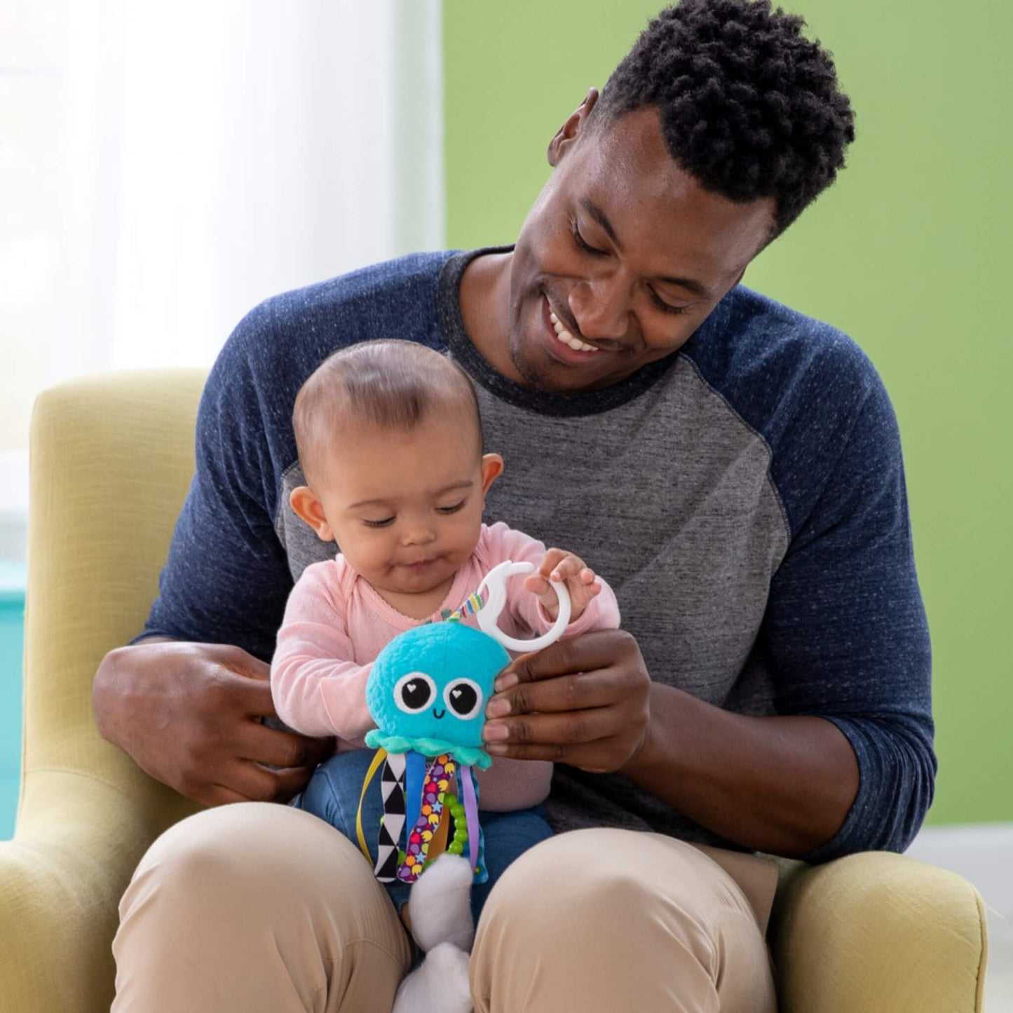 Father sitting indoors holding his baby, both engaged as the baby reaches for the jellyfish toy, exploring its ribbons and shapes during shared sensory play.