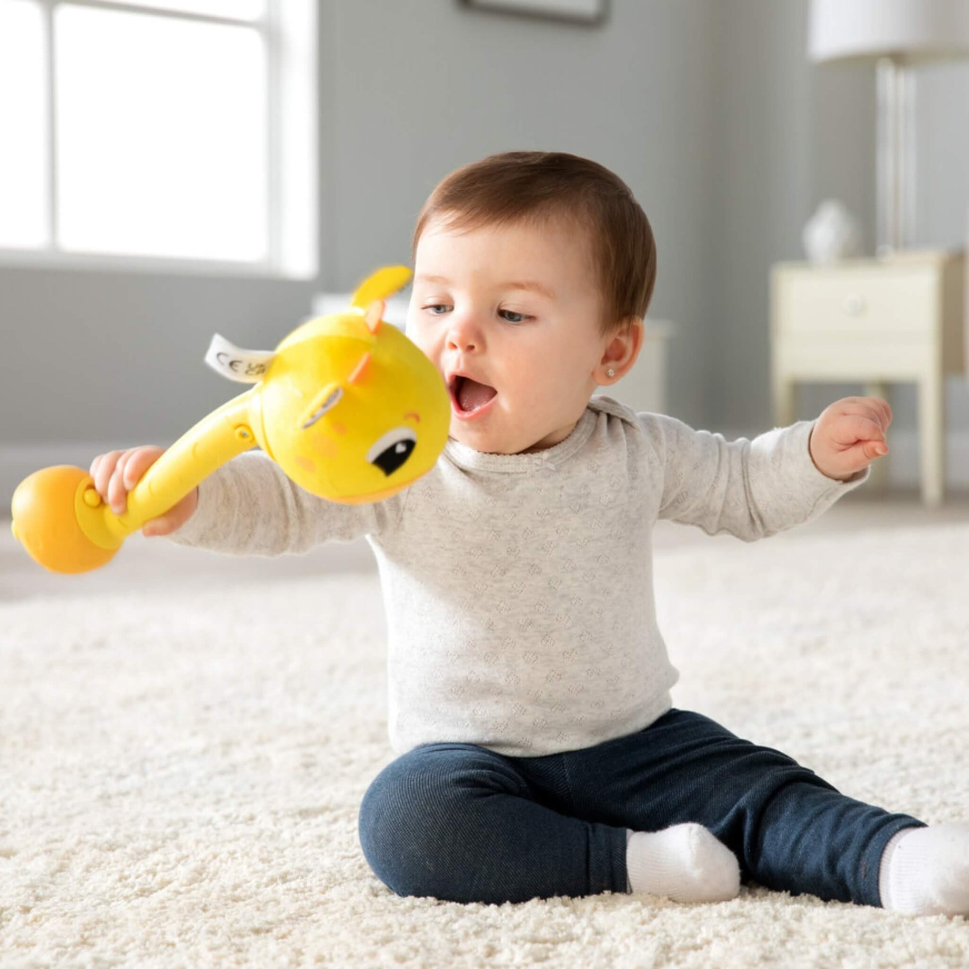 Baby sitting on a soft carpet holding a yellow giraffe rattle with a smiling face and plush head.