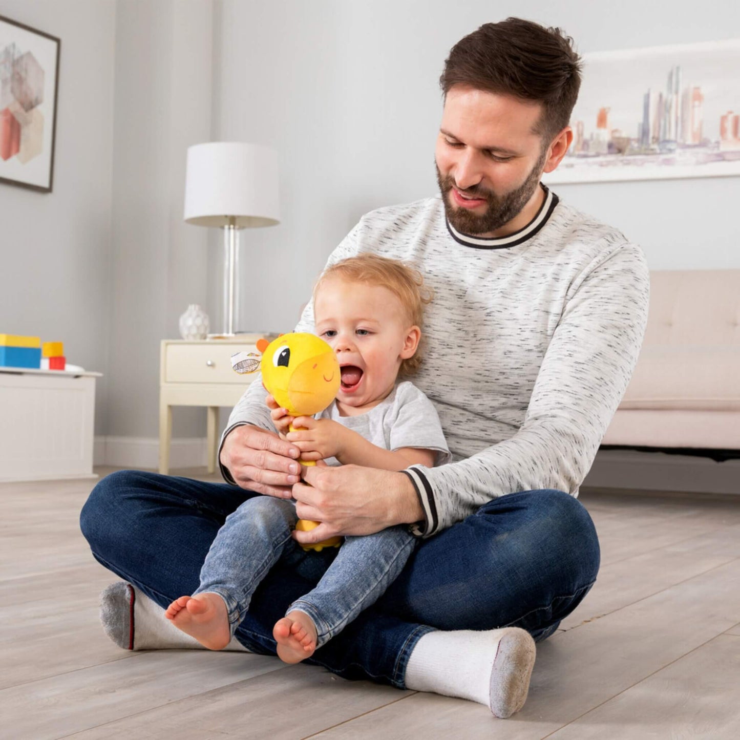 Father sitting cross-legged on a wooden floor with a toddler on his lap playing with a yellow giraffe baby toy.