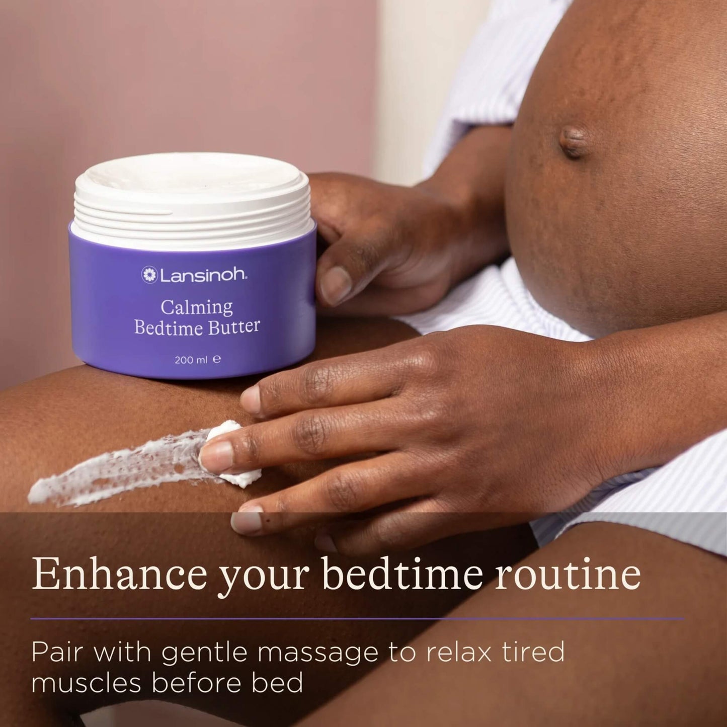 A pregnant woman sits with a jar of body butter beside her bump while applying a layer of cream to her leg.