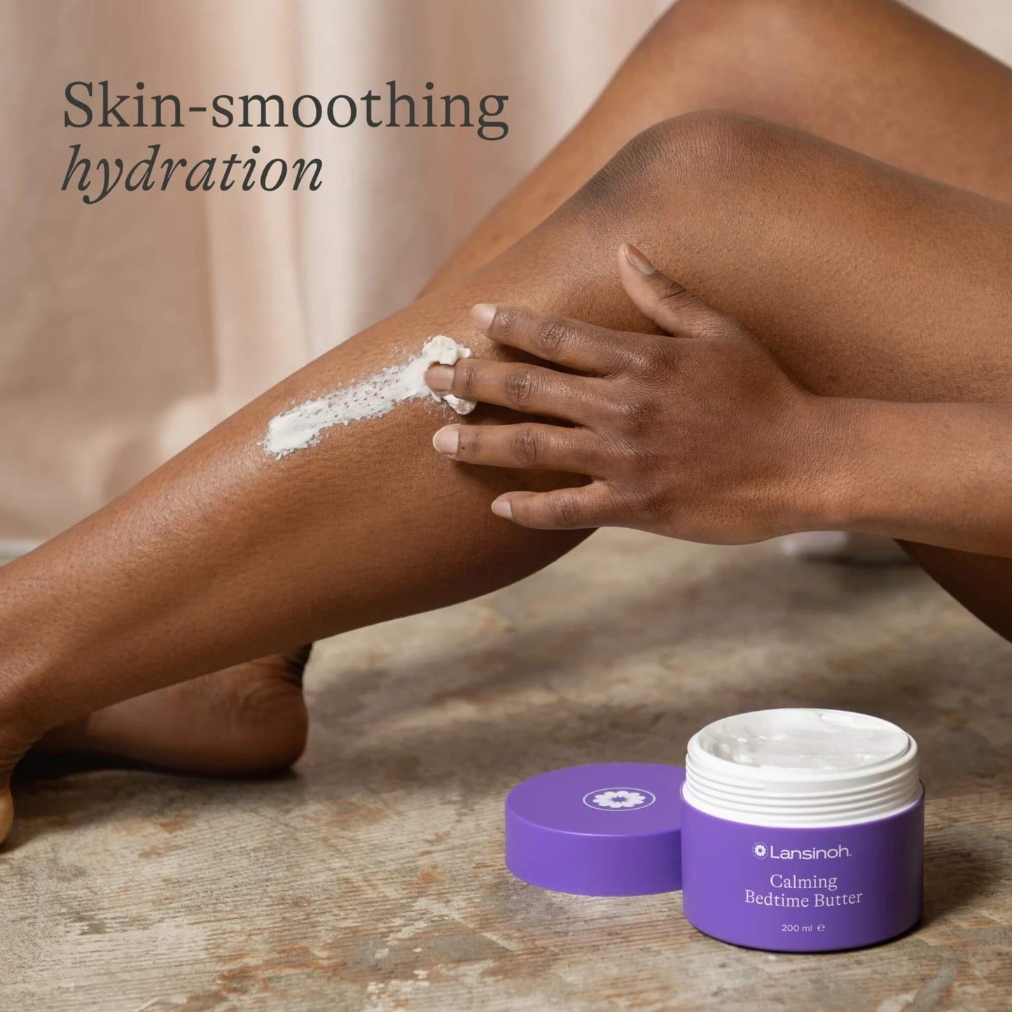 A woman massages white body butter into her lower leg while an open purple tub and lid rest on a wooden floor nearby.