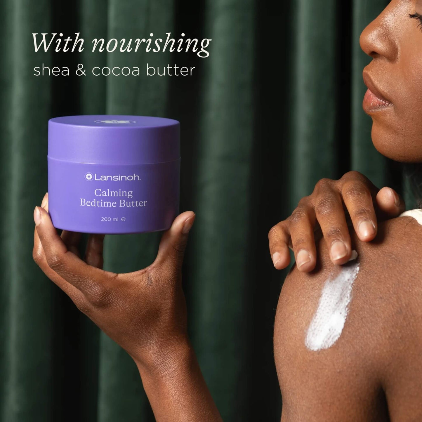 A woman holds a purple tub of body butter while applying cream to her shoulder against a dark green curtain backdrop.