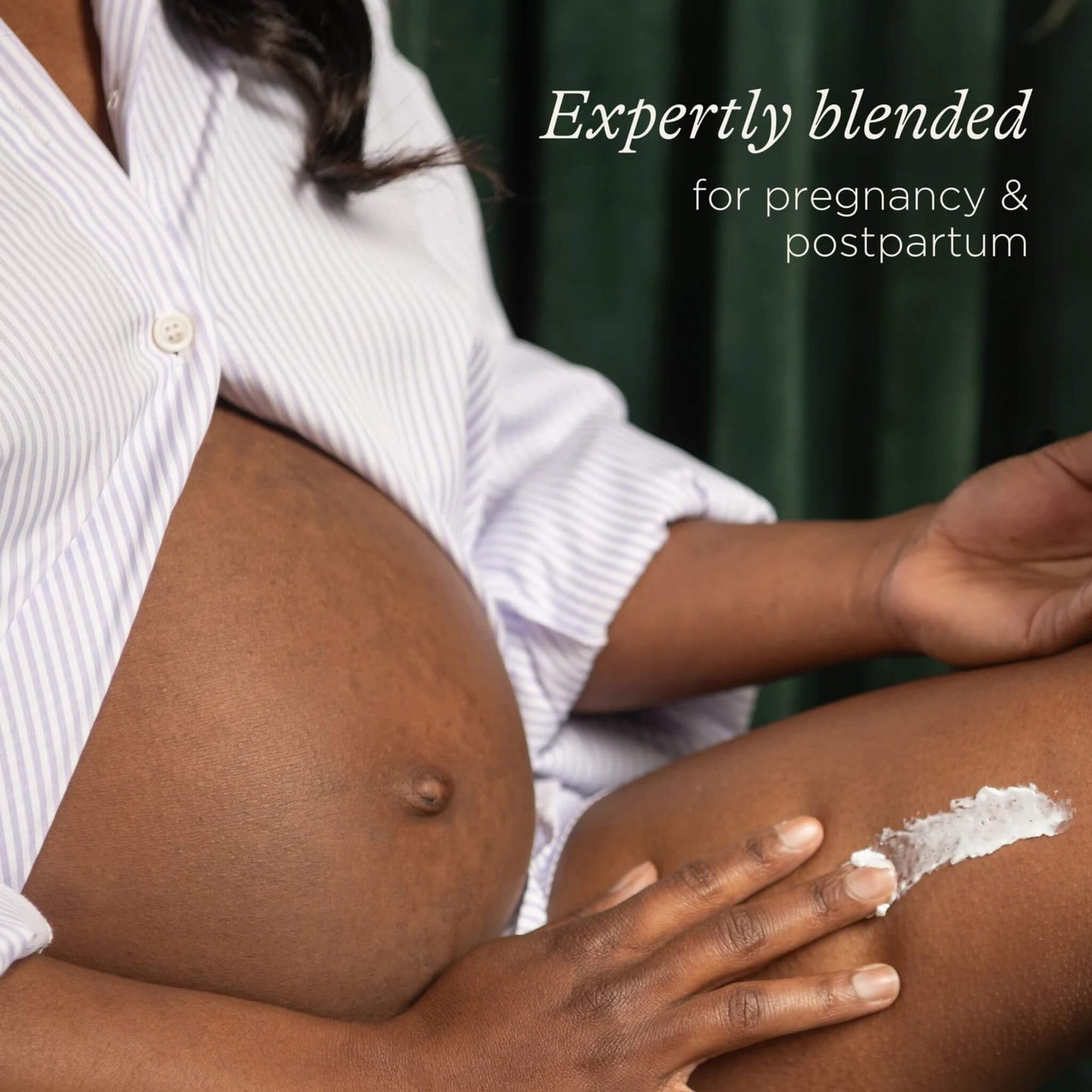 A pregnant woman applies body butter to her leg while sitting down, with her baby bump visible and a striped shirt draped loosely over her shoulders.