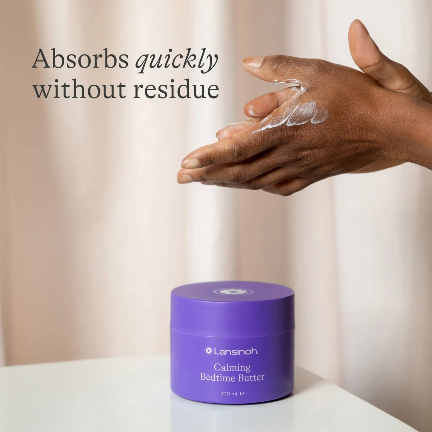 A pair of hands rubs white body butter into the skin above a purple tub placed on a white table.