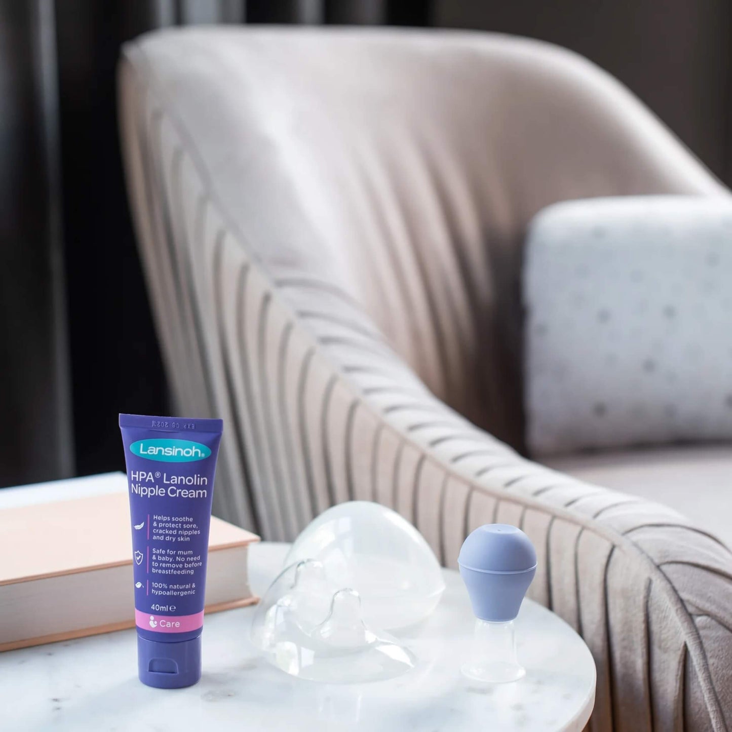 Transparent nipple shields with protective case and nipple care accessories arranged on a side table beside an upholstered chair.