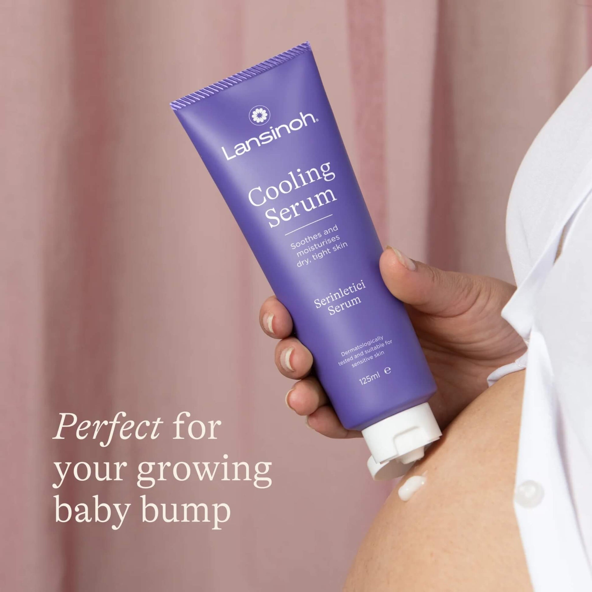 A woman squeezes cooling serum onto her pregnant belly from a purple tube against a soft pink backdrop.