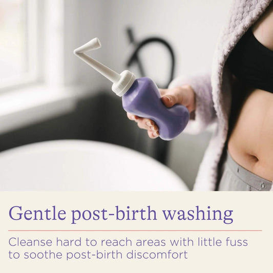 A woman holding a purple post-birth wash bottle beside a bath, showing the angled spout designed to help cleanse hard-to-reach areas after childbirth.