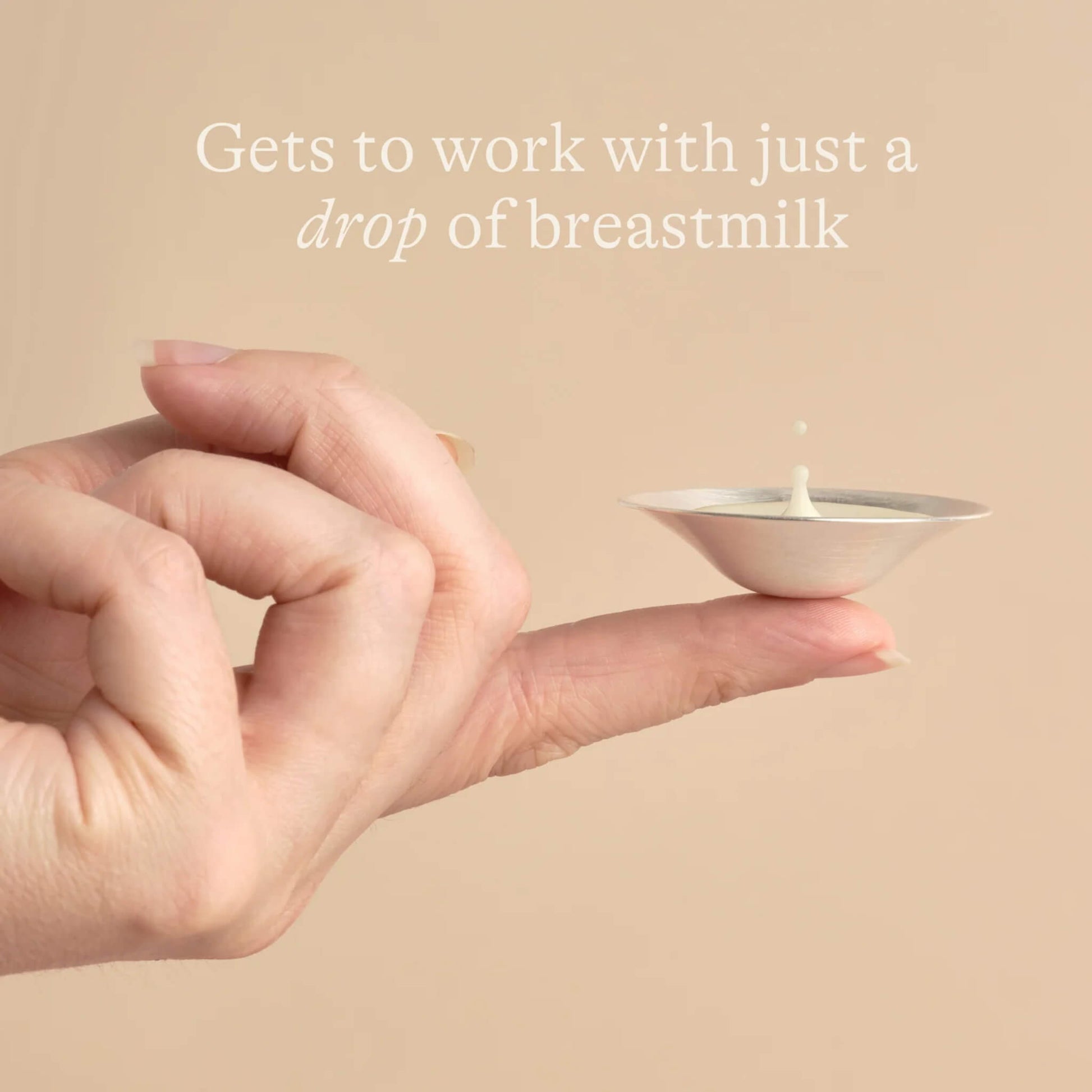 Close-up of hand balancing a silver nipple cup on one finger with a drop of breast milk inside, illustrating how only a small amount of milk is needed for soothing use.