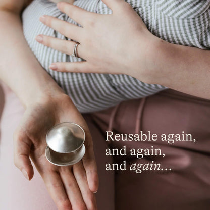 Mother resting a pair of silver nipple cups in her hand while cradling her baby, showing the reusable and durable nature of the Silverette design.