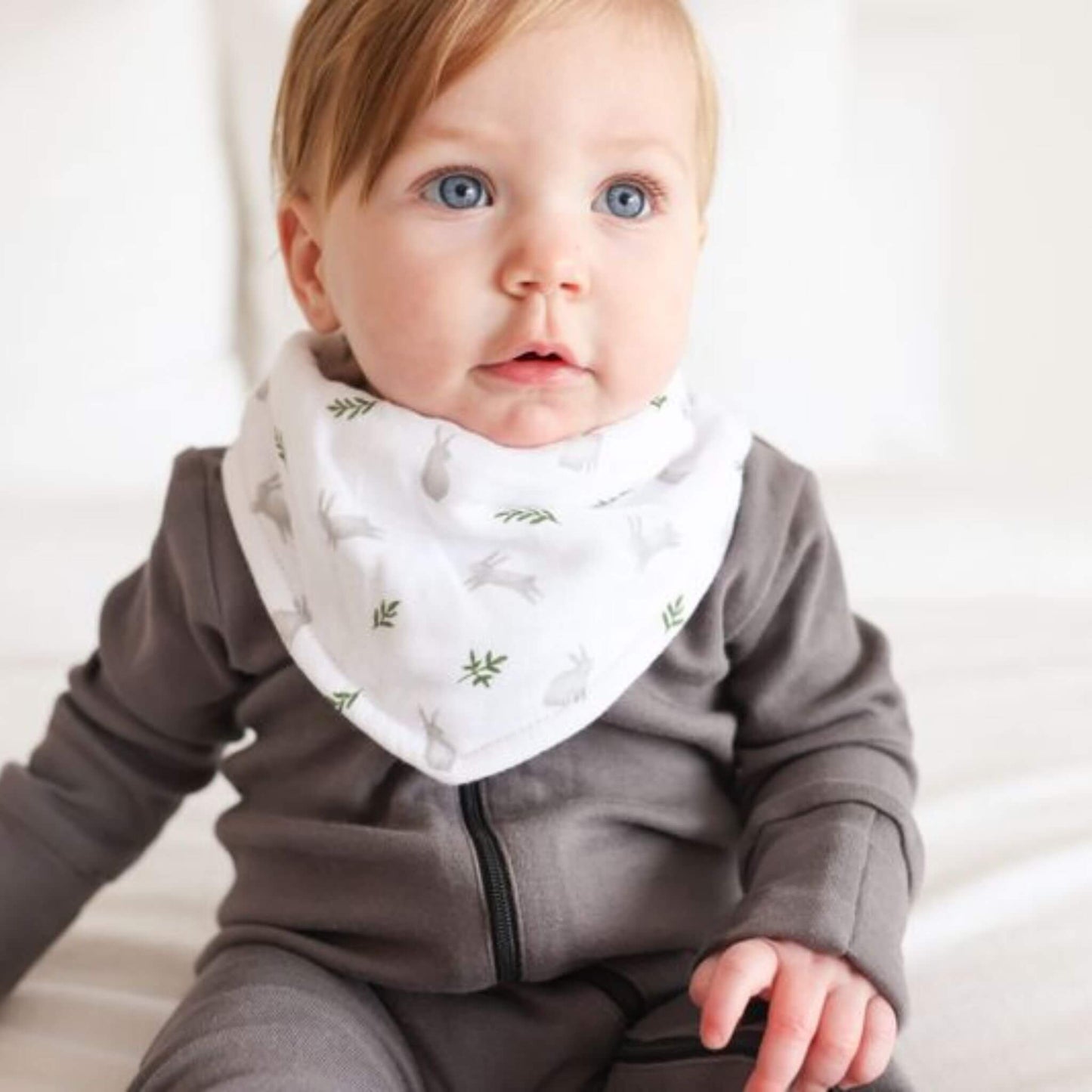 Baby with bright blue eyes wearing a sage green Lulujo bandana bib whilst sitting comfortably on white bedding, showcasing the perfect fit and earthy colour palette.