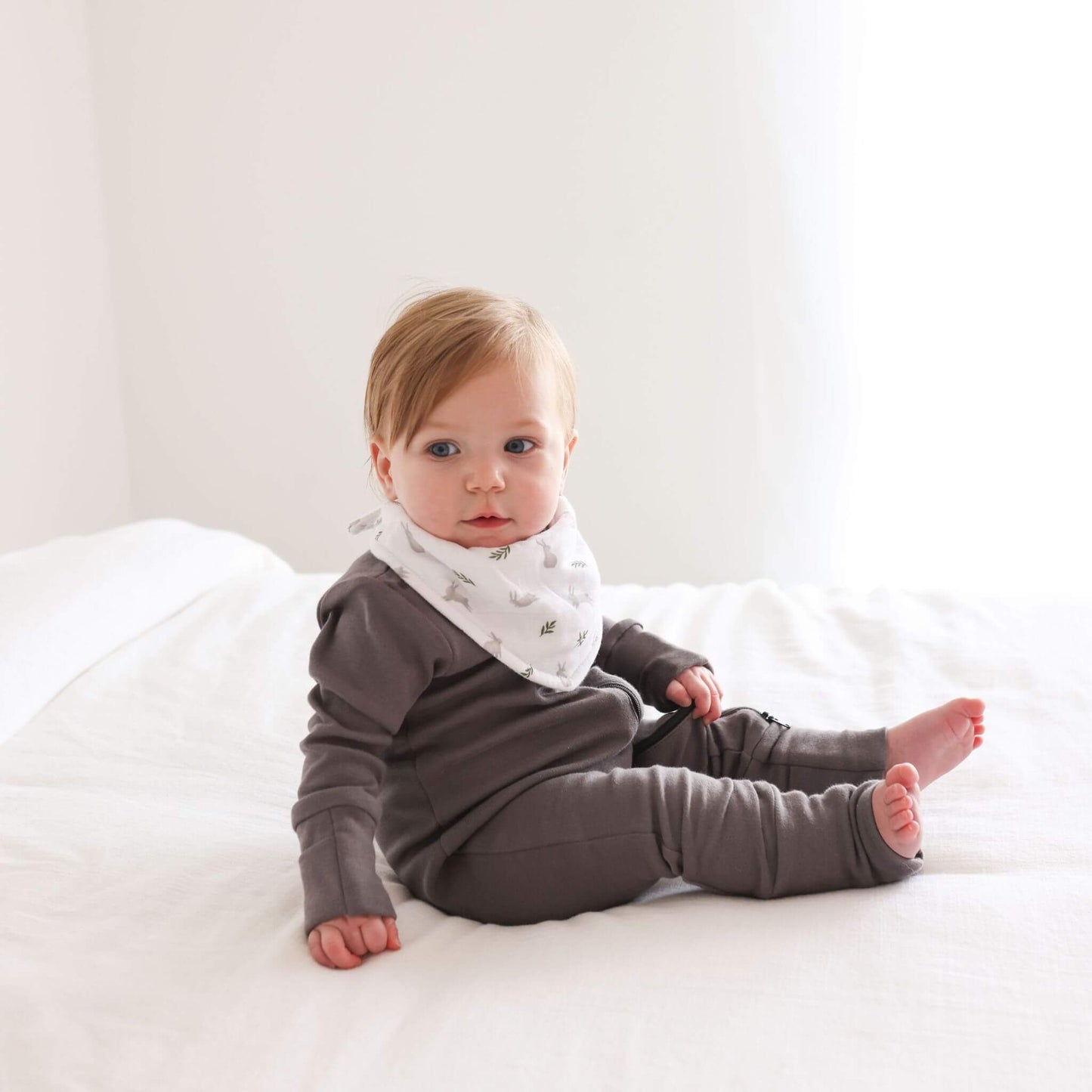 Close-up of baby wearing white Lulujo bandana bib with bunny and fern pattern, displaying the soft fabric against delicate skin and stylish grey outfit coordination.