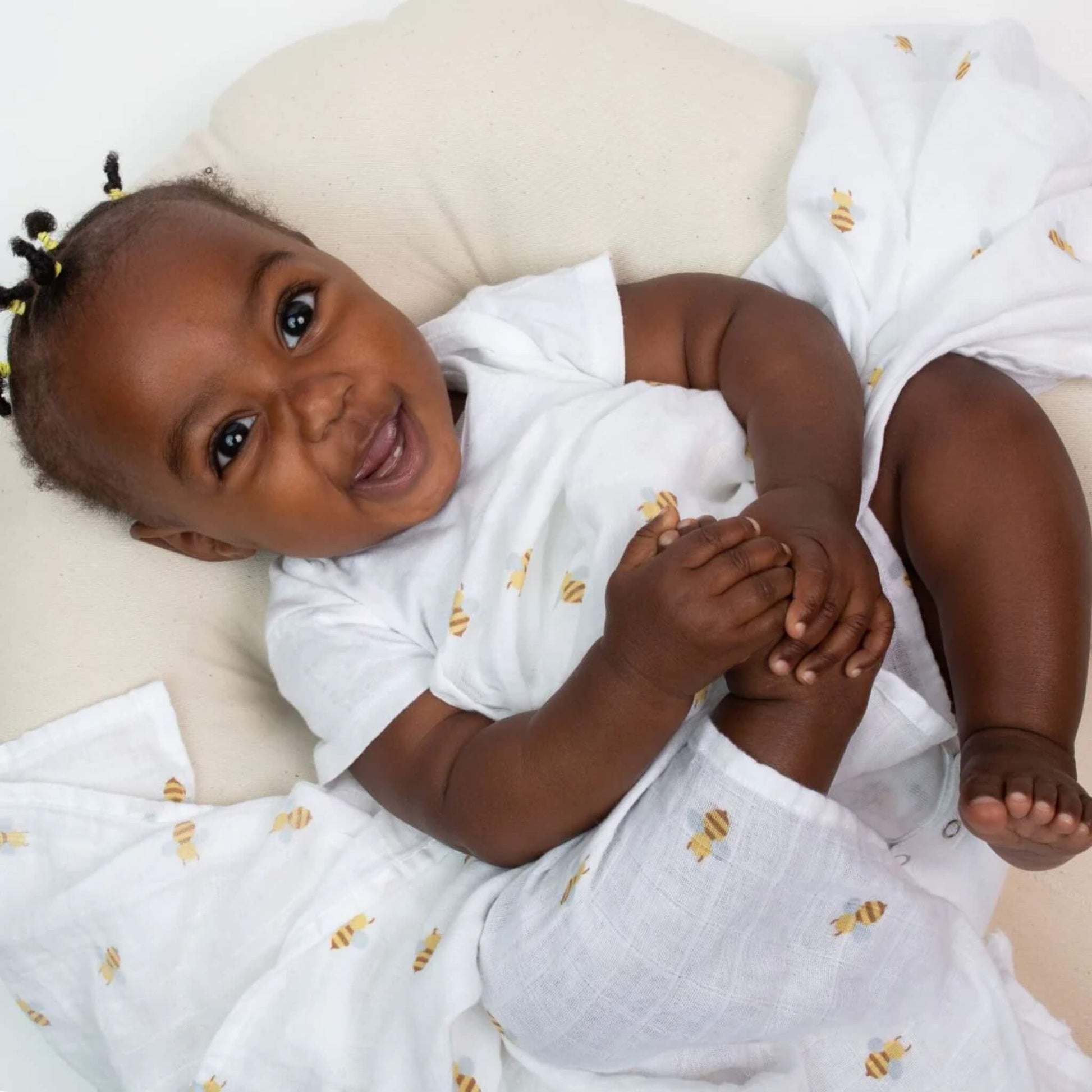 Smiling baby wrapped in a white muslin swaddle with yellow bee pattern, lying comfortably on a soft cream cushion.