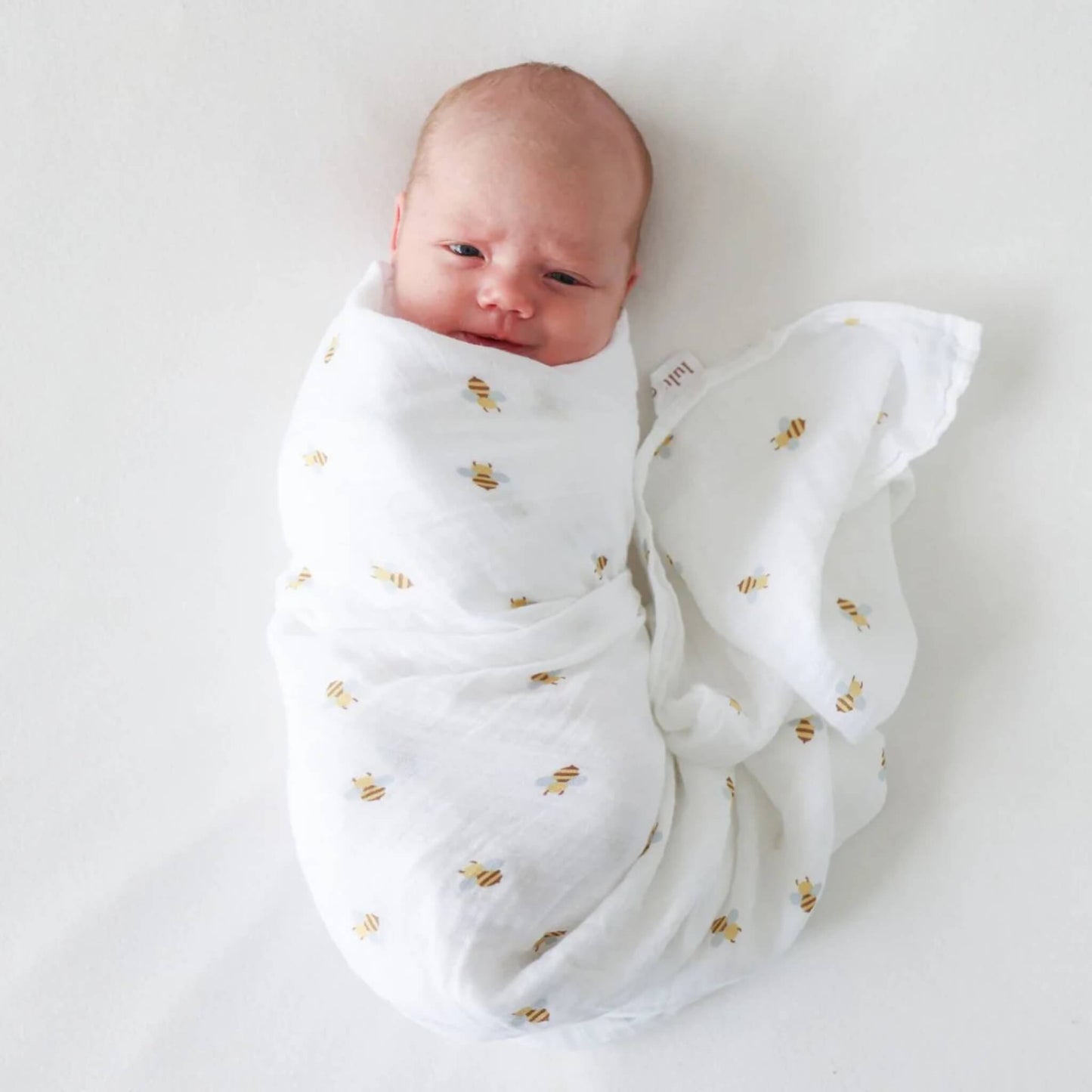 Newborn snugly wrapped in the bee-print muslin swaddle, resting on a white background.