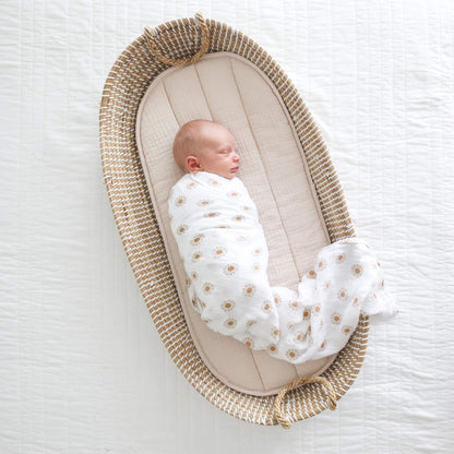 Sleeping newborn wrapped in patterned muslin and placed in a woven Moses basket on a quilted white surface.