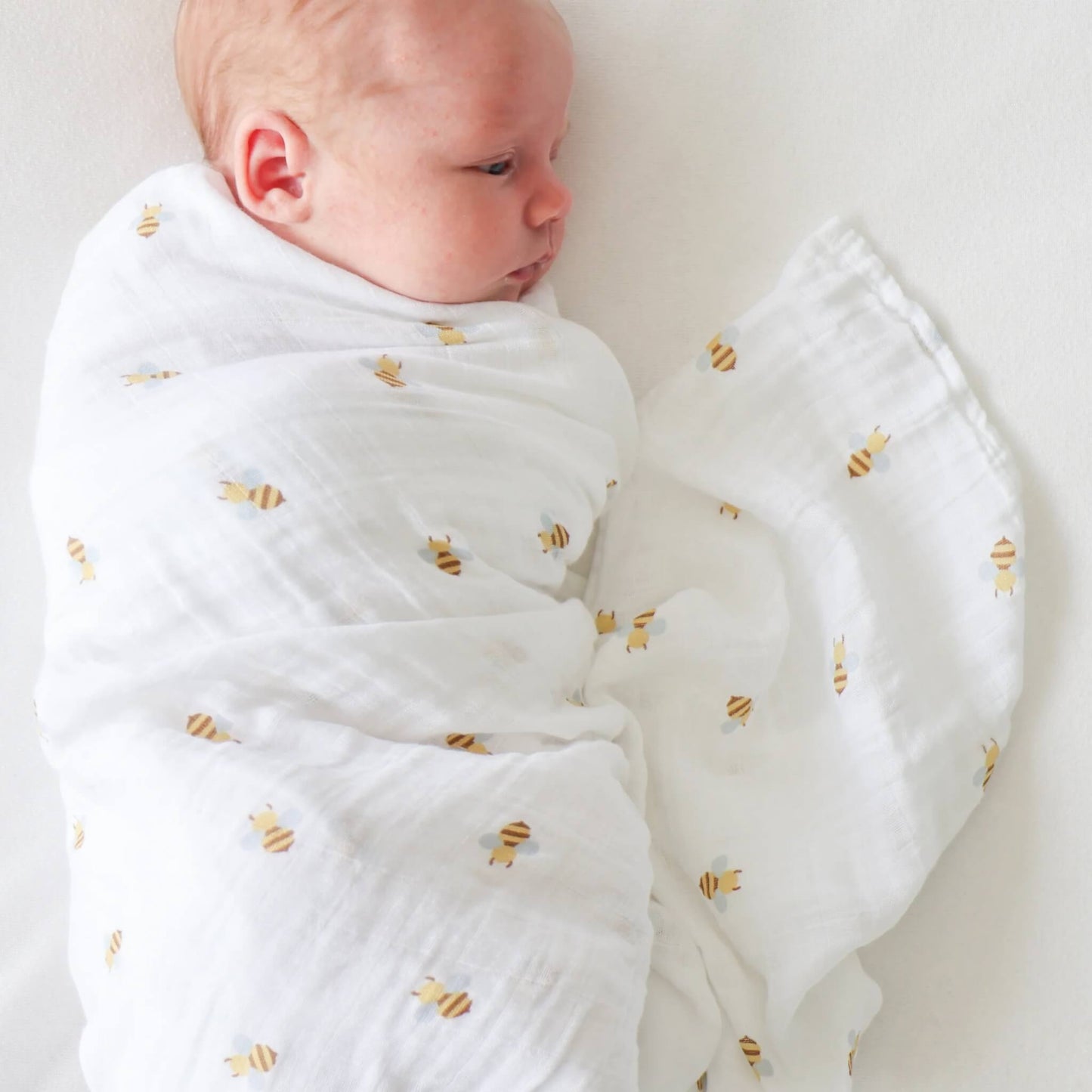 Baby swaddled in the white cotton muslin with small yellow bees, lying on their side with fabric softly gathered around.