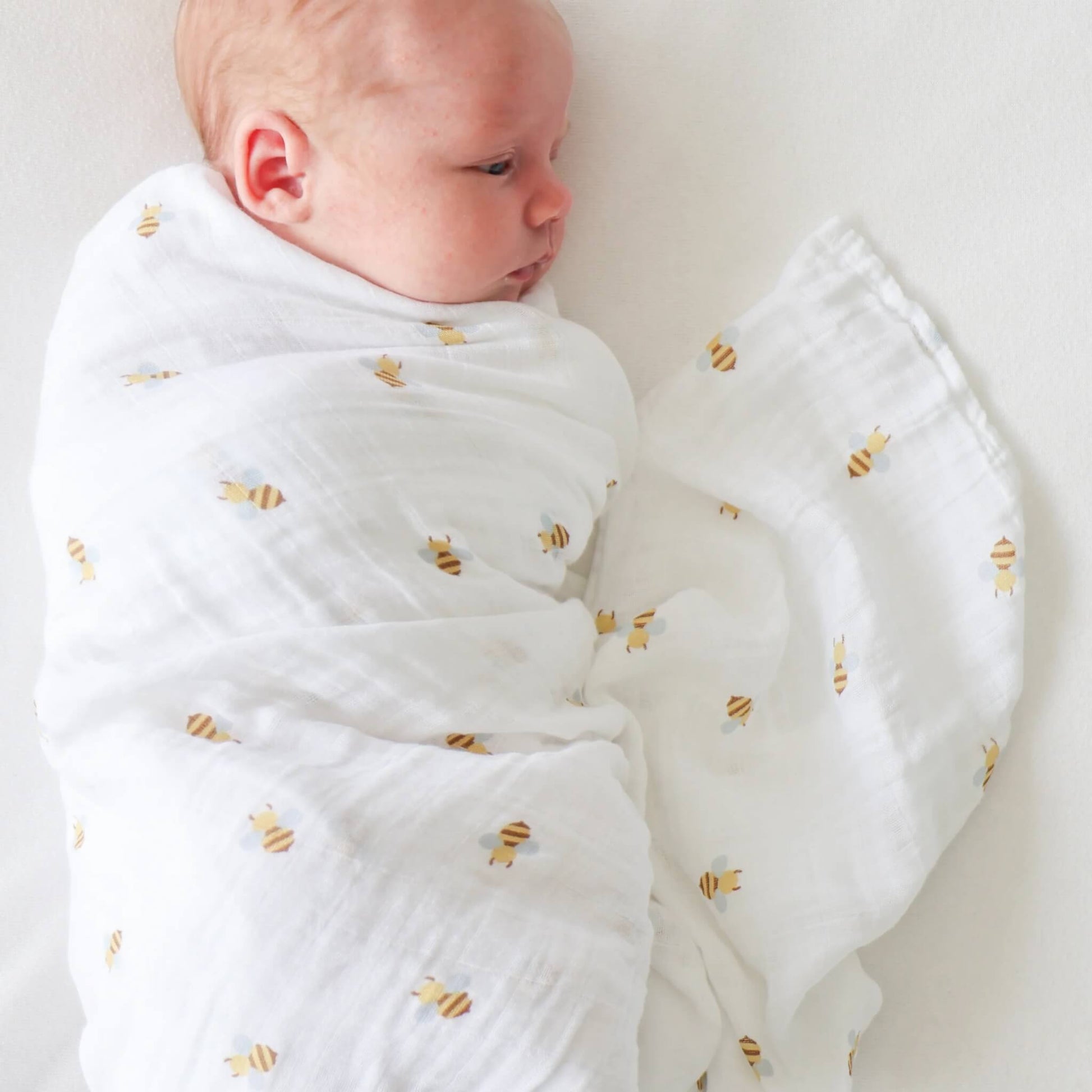Baby swaddled in the white cotton muslin with small yellow bees, lying on their side with fabric softly gathered around.