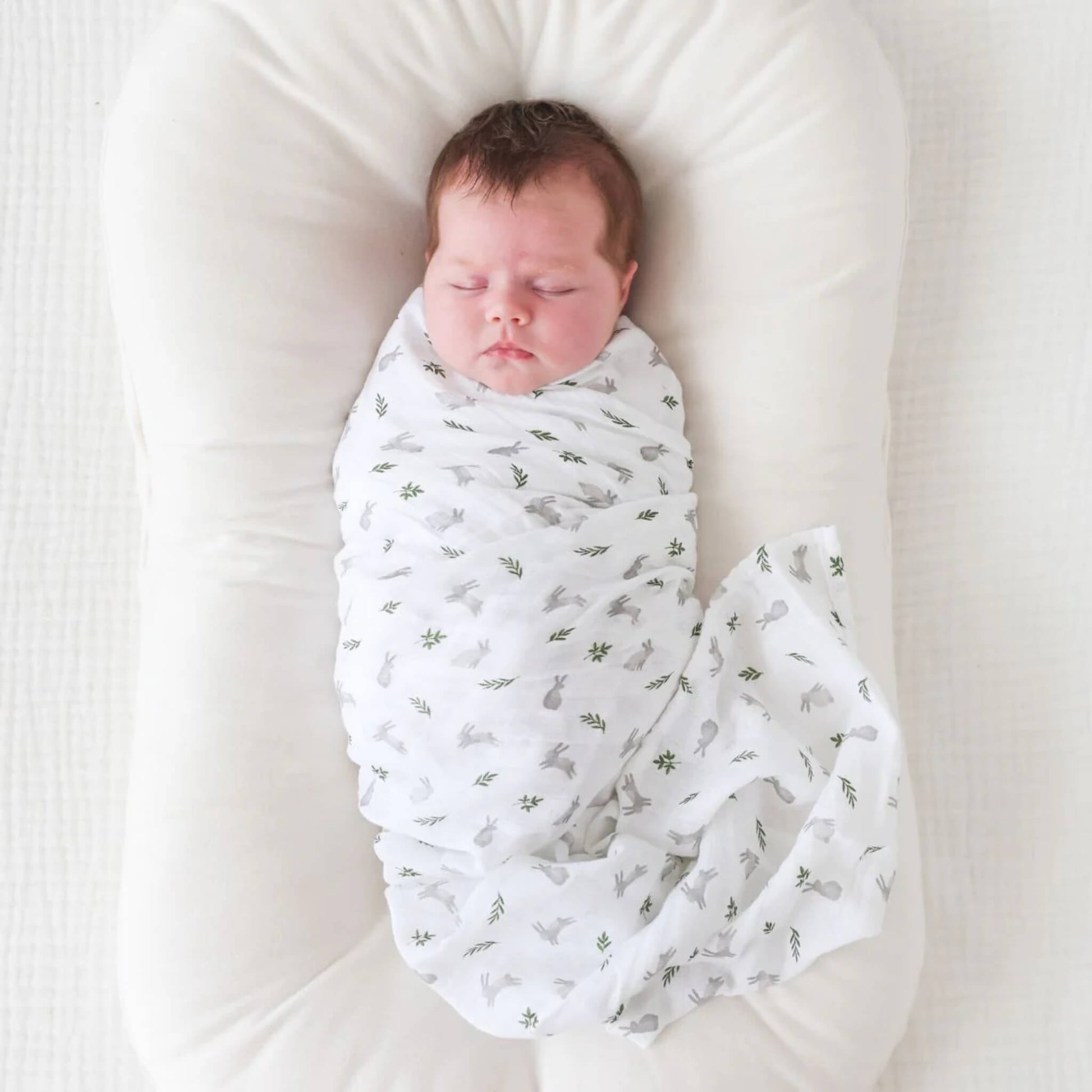 Newborn baby peacefully wrapped in the white muslin swaddle patterned with small bunnies and leaves, resting in a soft cushioned nest.