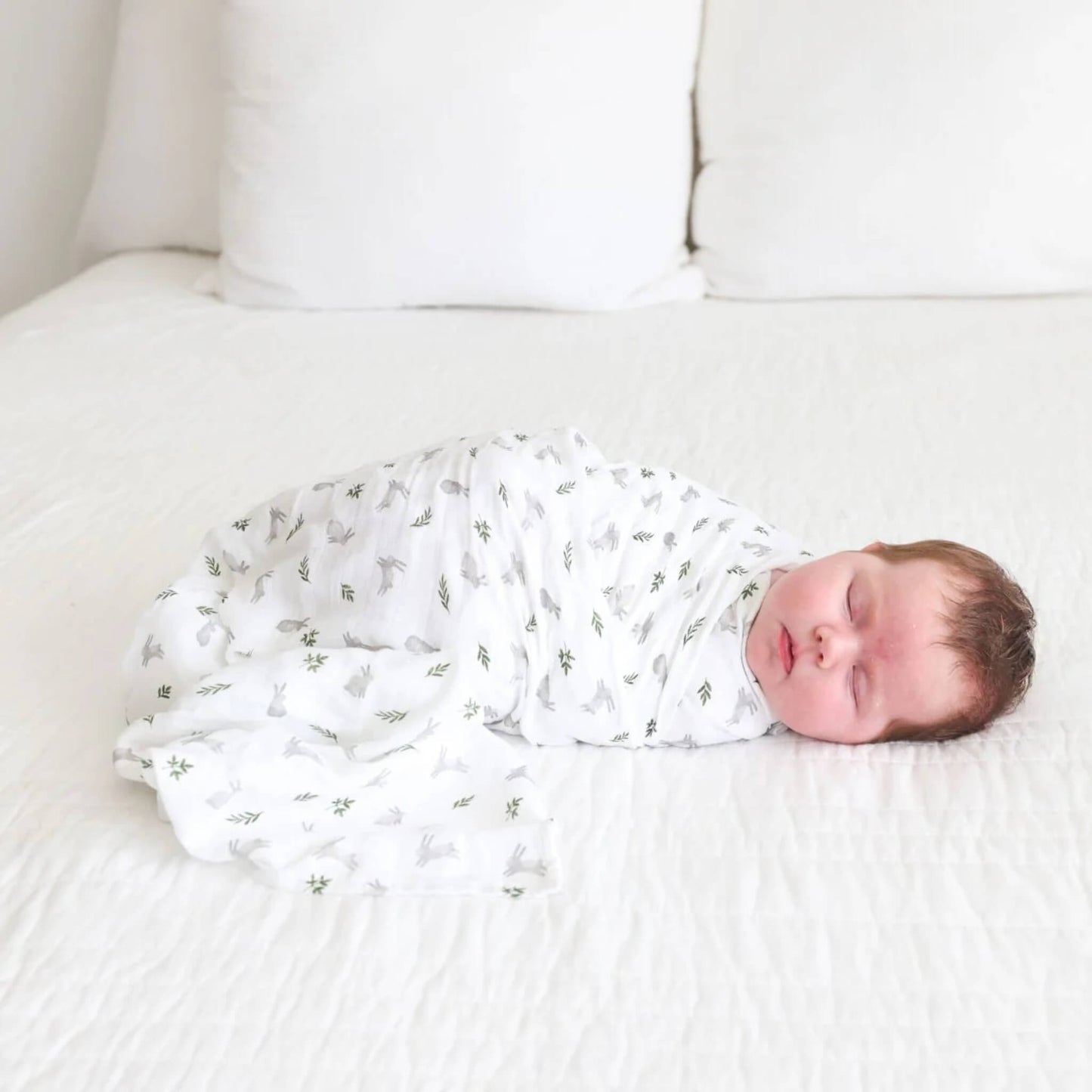 Baby lying asleep on a white bed, wrapped in the lightweight muslin swaddle featuring grey bunny illustrations.