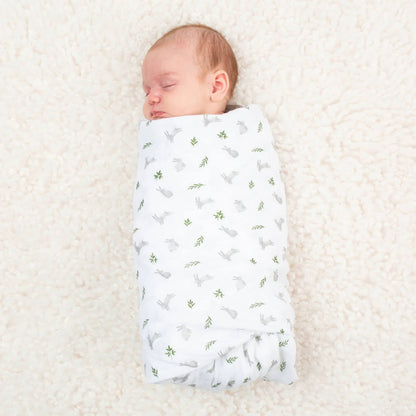 Newborn sleeping on a soft cream blanket, swaddled in a white muslin with a scattered bunny and leaf print.