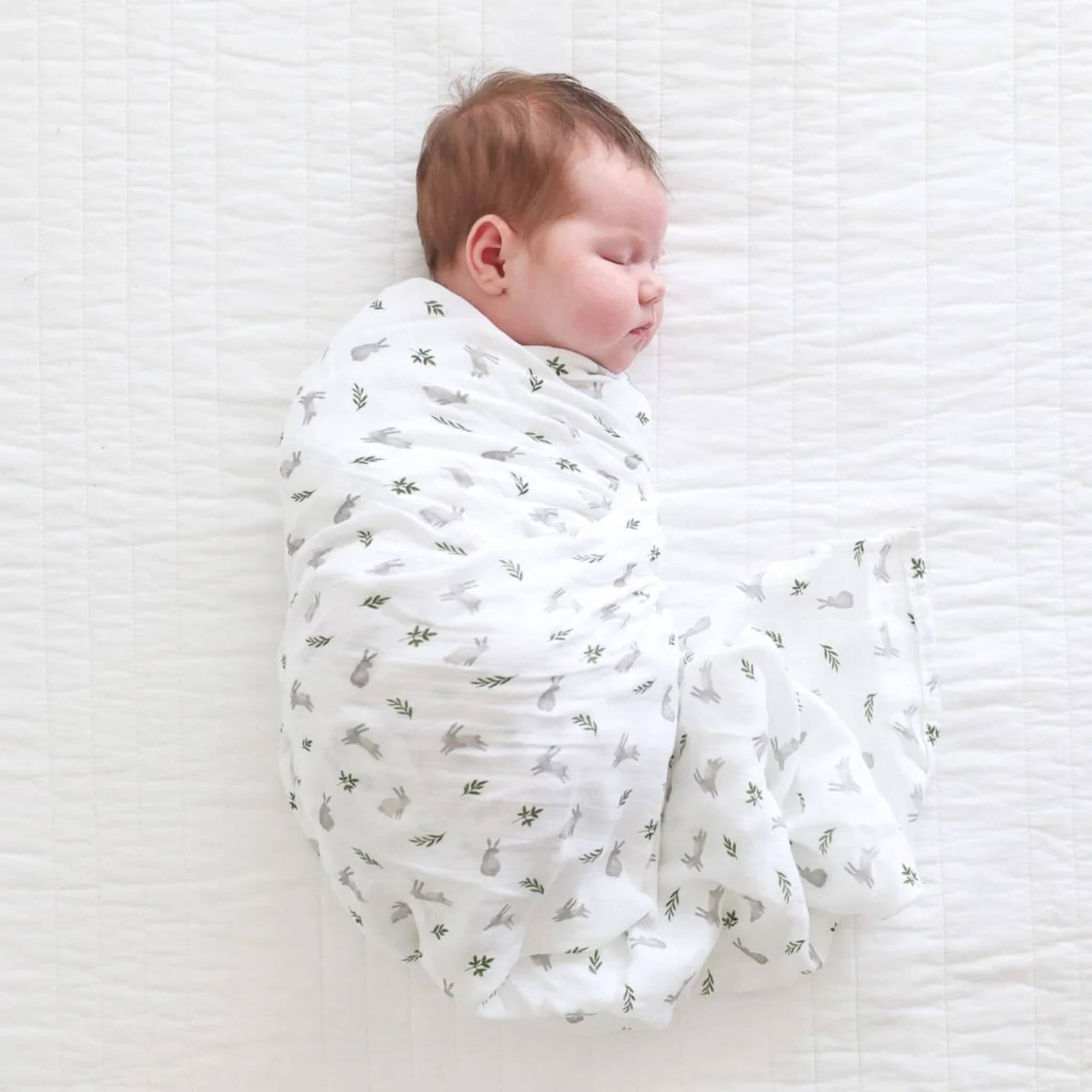 Baby swaddled in white muslin with grey bunnies and green leaves, resting on a lightly quilted surface.