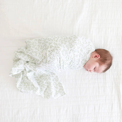 Newborn baby lying on a white bed, wrapped in the white muslin swaddle decorated with small green leaves.