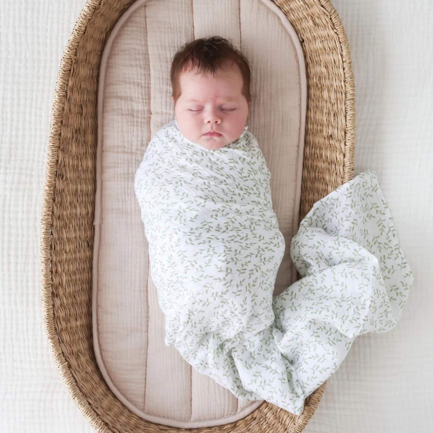 Baby sleeping peacefully in a woven Moses basket, swaddled in the white muslin with a soft green leaf pattern.
