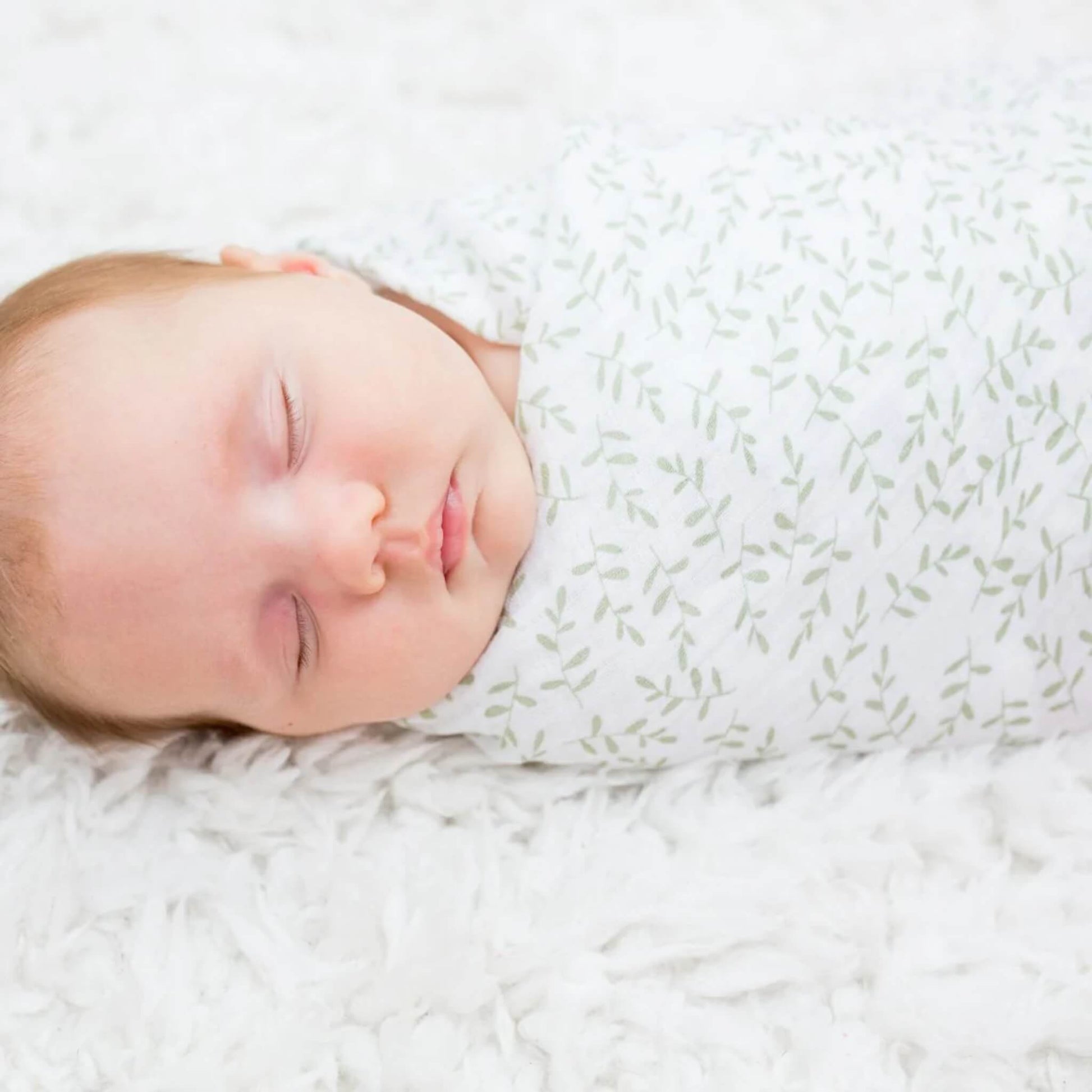 Close-up of a sleeping baby swaddled in white muslin patterned with delicate green leaves, lying on a soft white textured blanket.