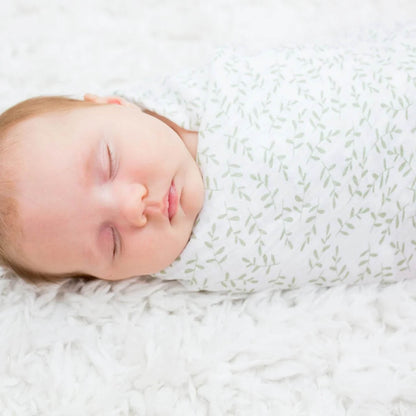 Close-up of a sleeping baby swaddled in white muslin patterned with delicate green leaves, lying on a soft white textured blanket.