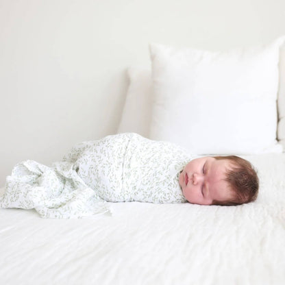 Baby resting on a white bed, swaddled in the lightweight muslin fabric printed with pale green leaves.