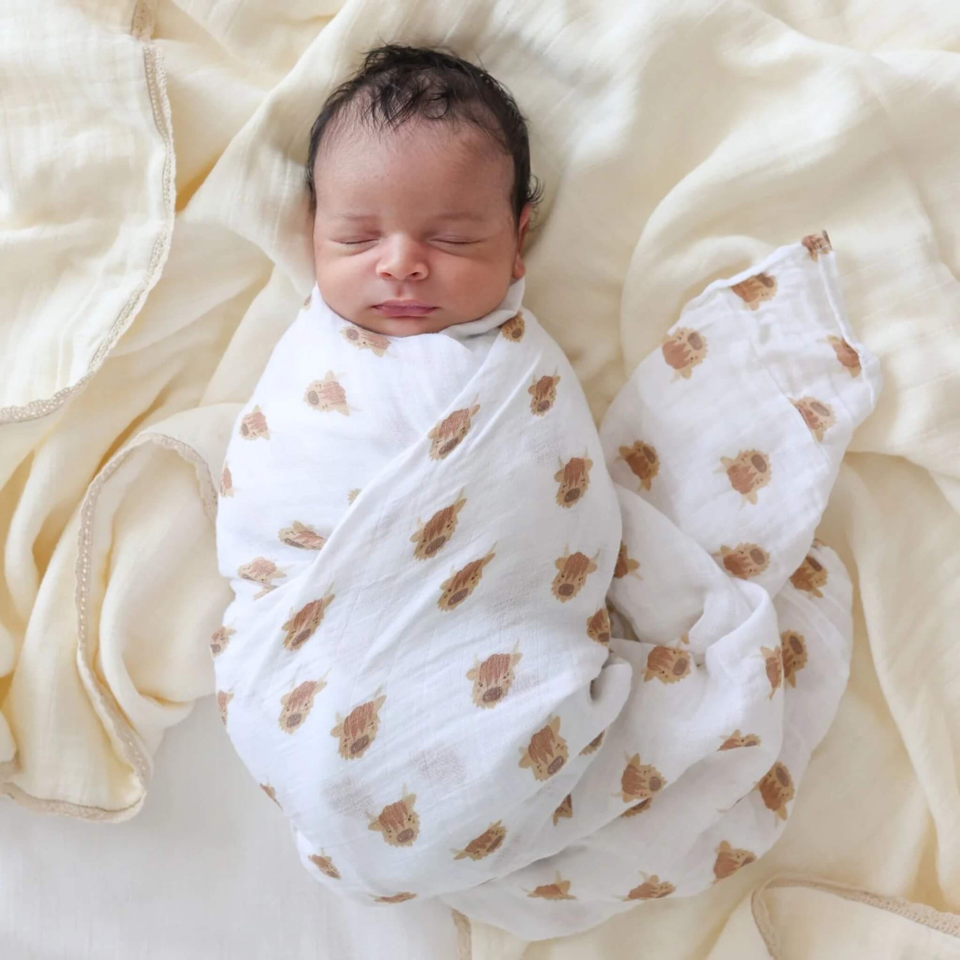 Newborn baby wrapped in the white muslin swaddle covered with brown Highland cow faces, resting on soft cream bedding.