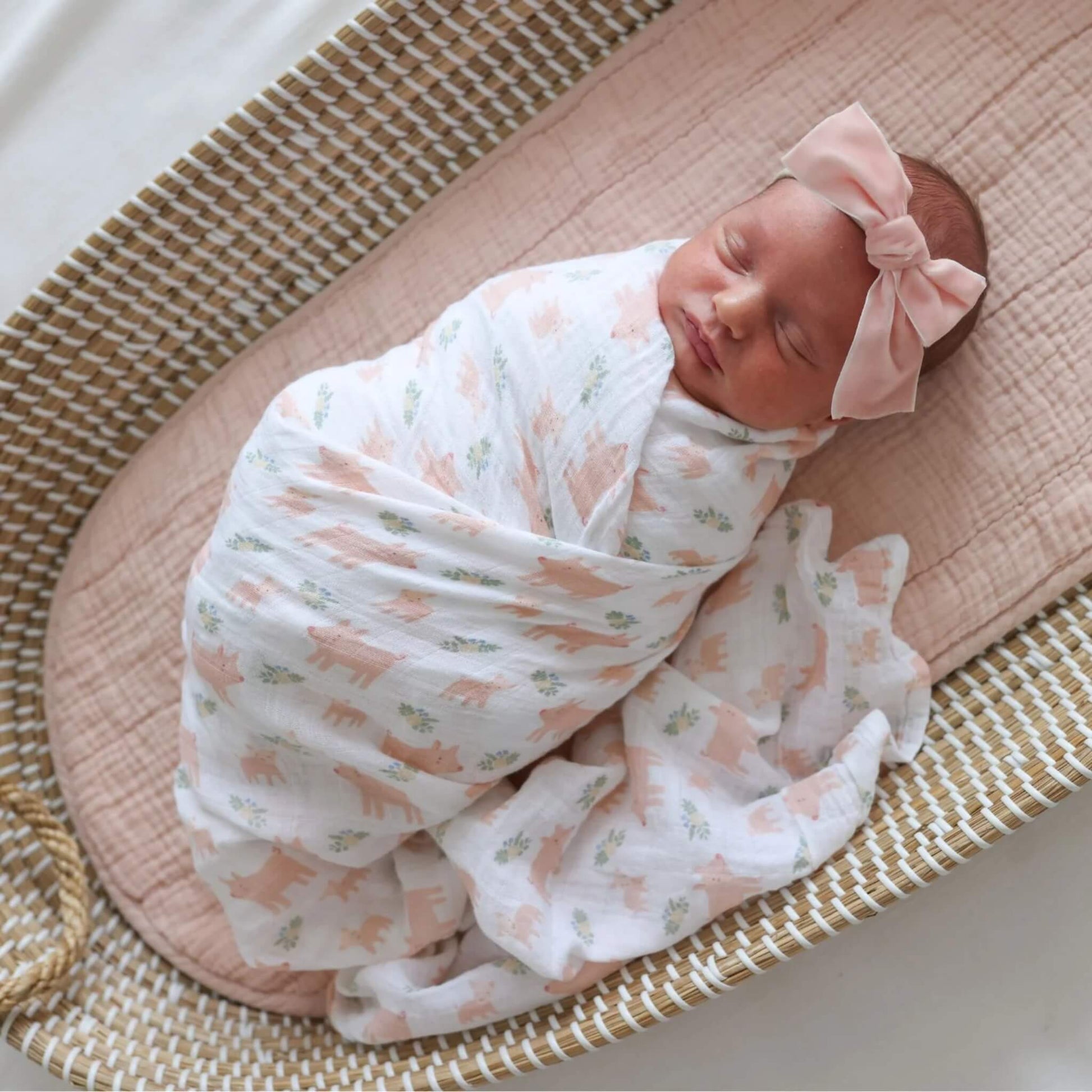 Newborn baby sleeping in a woven Moses basket, swaddled in the white muslin blanket printed with little pink pigs.