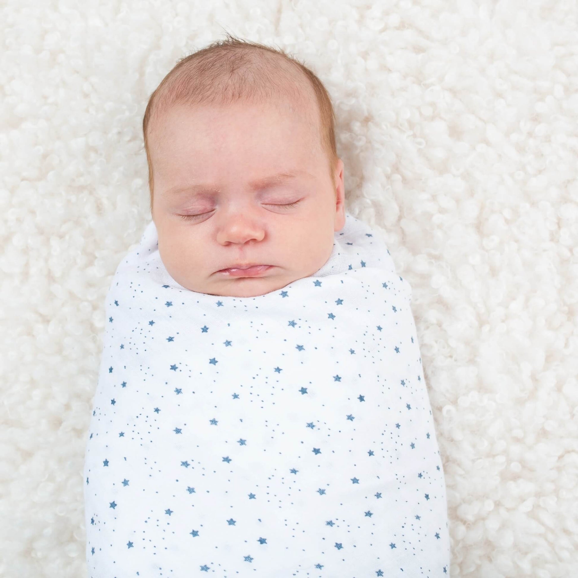 Baby peacefully sleeping, wrapped in the white muslin blanket covered with tiny blue stars on a soft textured surface.