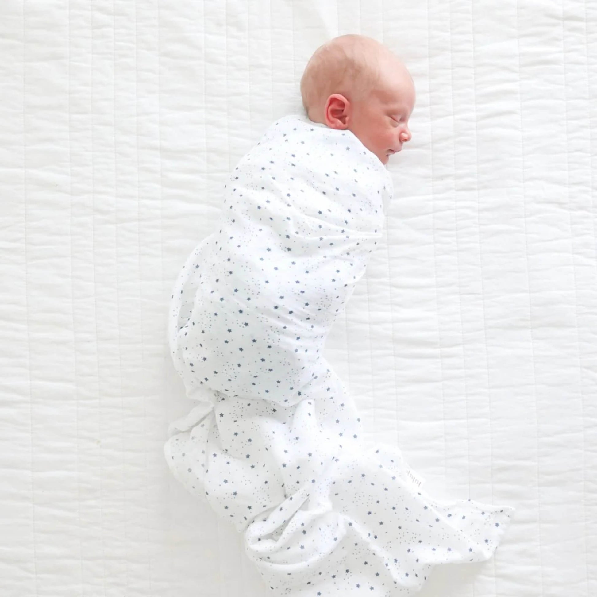 Side view of a newborn wrapped snugly in the star-patterned muslin swaddle, resting on a white quilt.