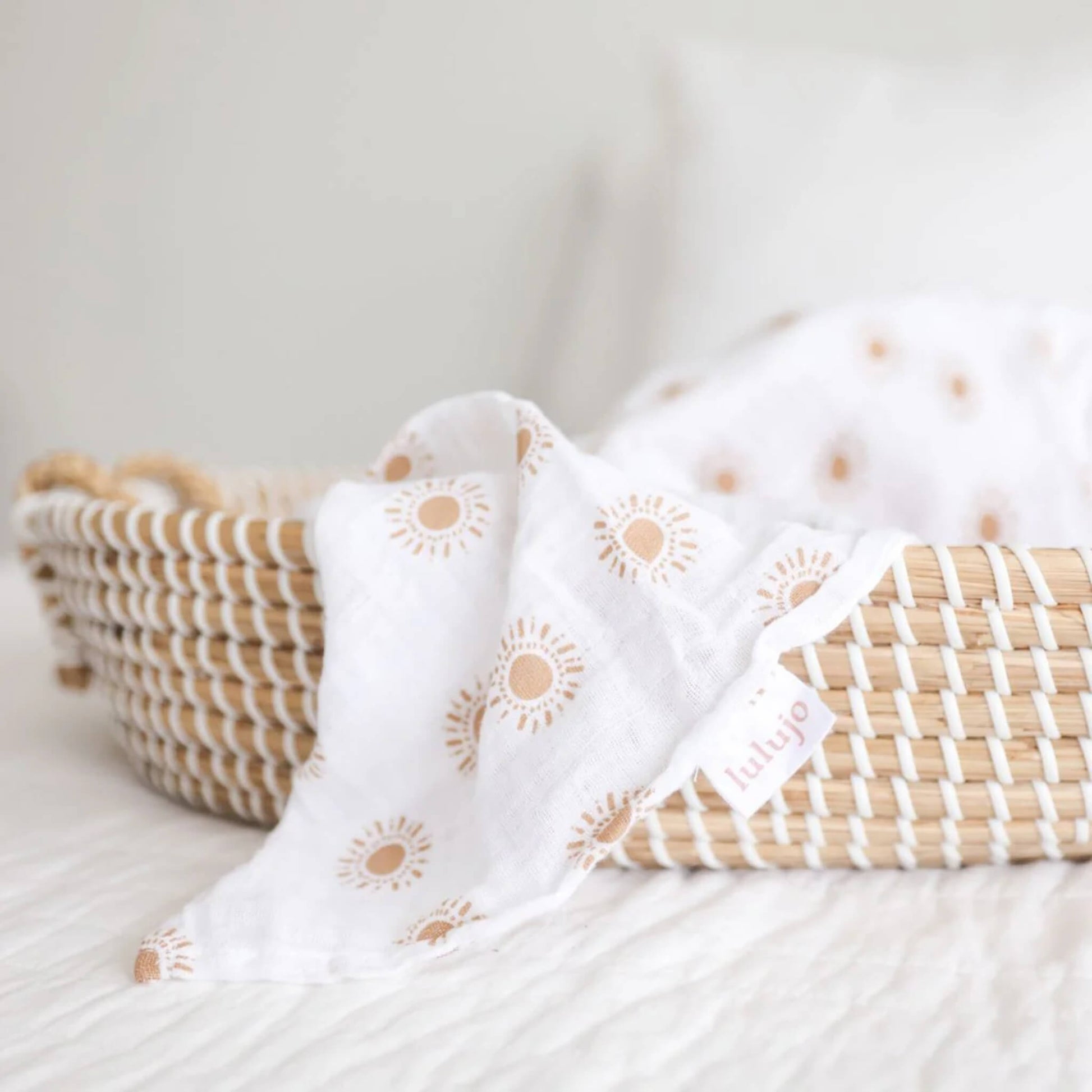Detail shot of a muslin swaddle draped over the edge of a woven Moses basket, showing the Lulujo fabric tag and sun print.