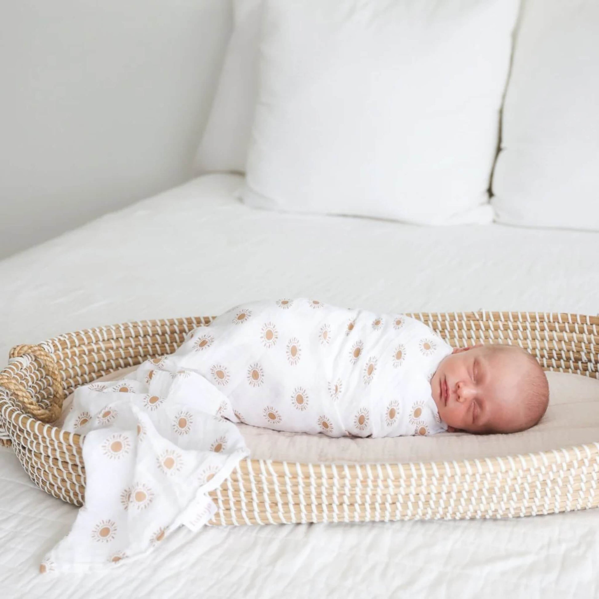 Baby sleeping in a woven Moses basket, wrapped snugly in white muslin swaddle printed with golden suns.