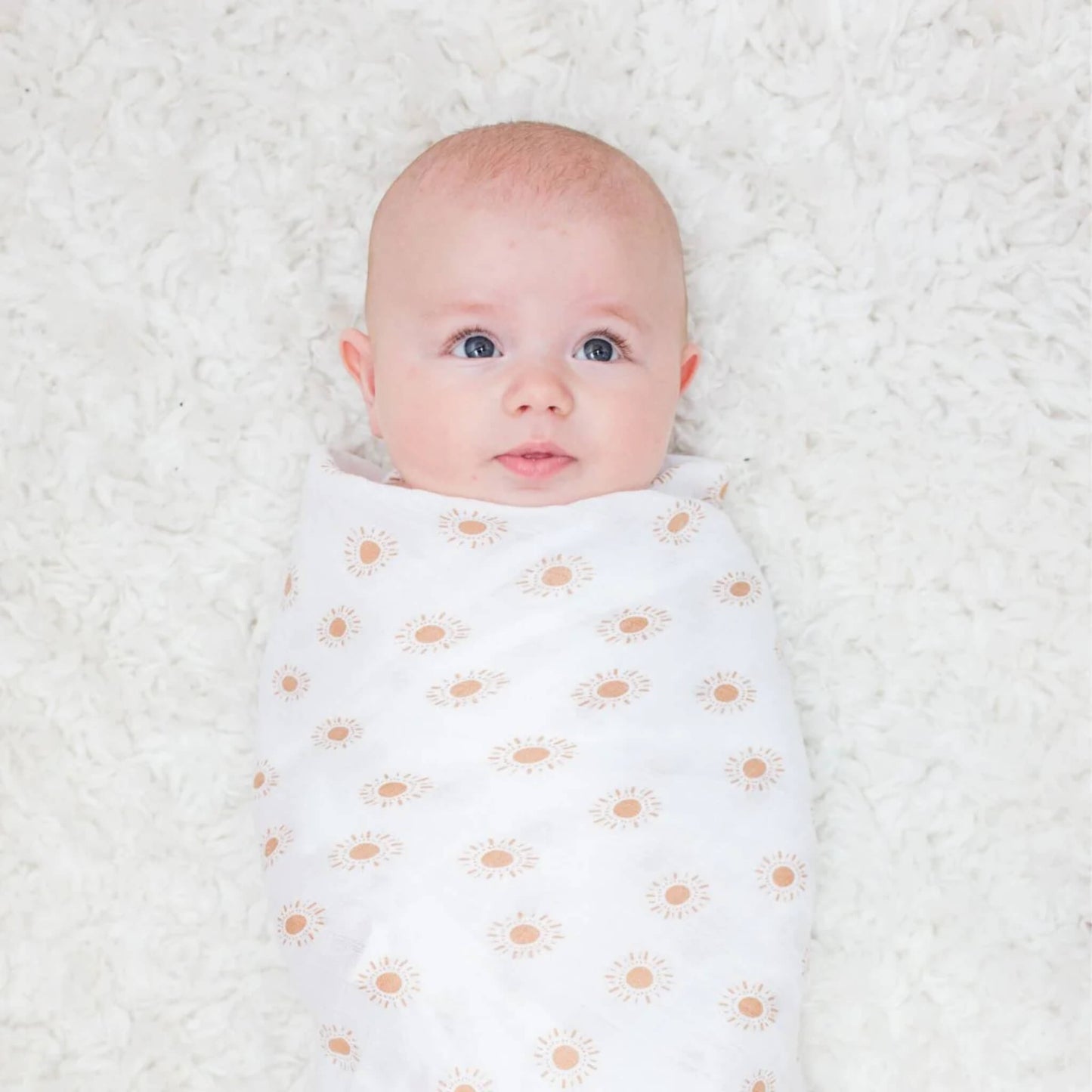 Baby lying on a soft white rug, swaddled in muslin blanket decorated with mustard-yellow suns.