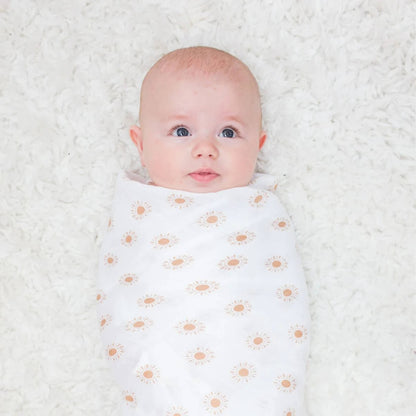 Baby lying on a soft white rug, swaddled in muslin blanket decorated with mustard-yellow suns.