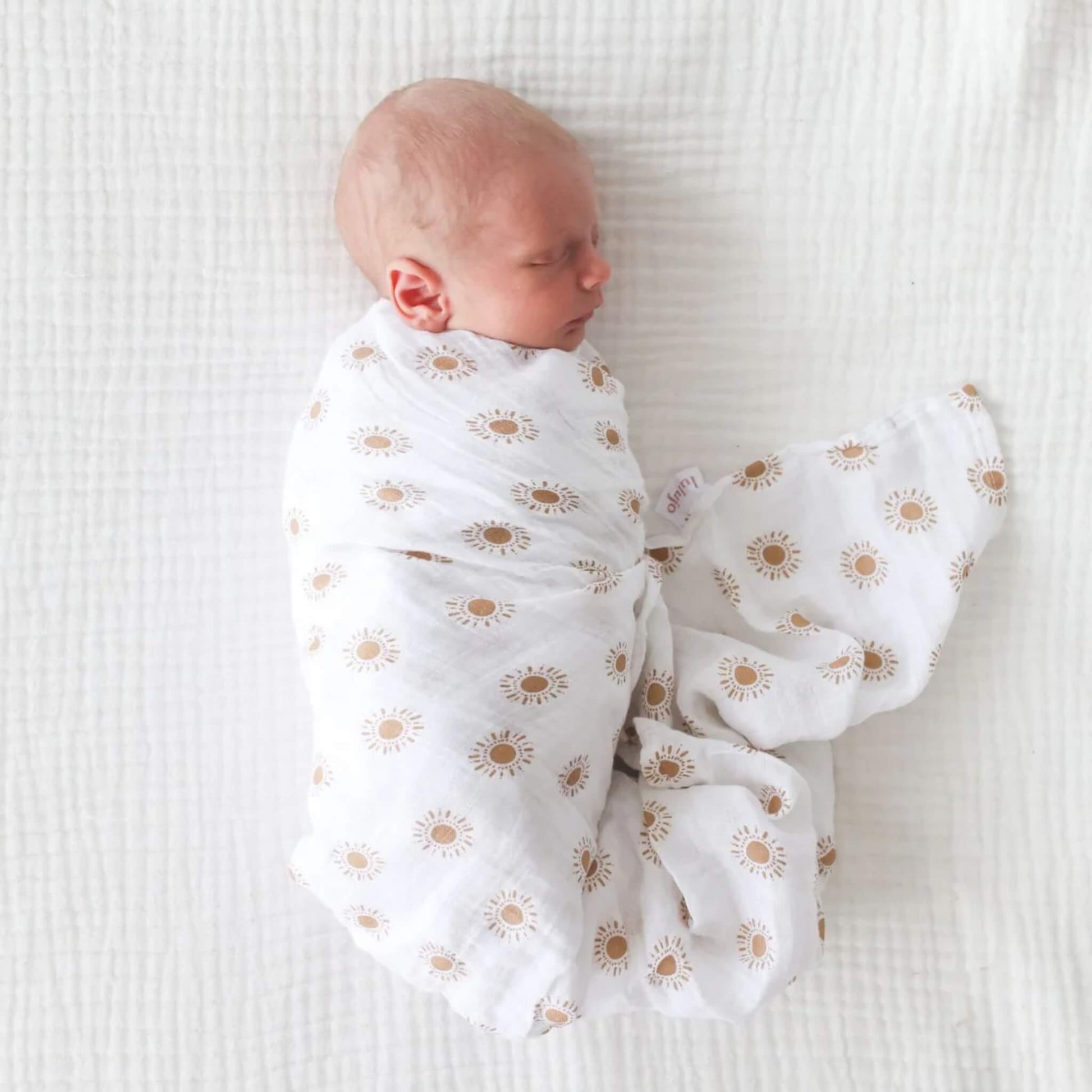 Side view of newborn sleeping soundly in sun-patterned muslin swaddle on white quilted bedding.
